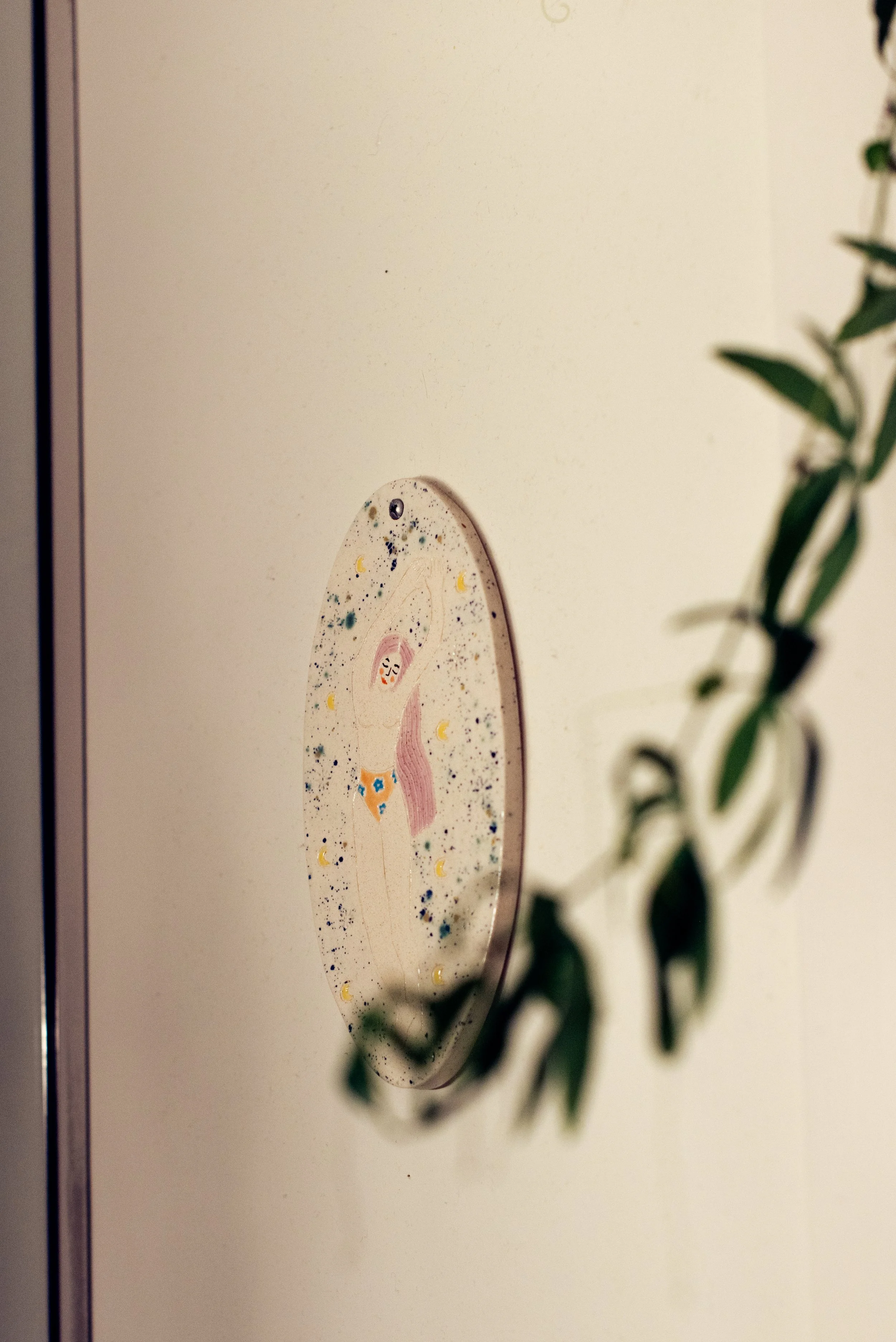 Decorative plate hanging on a white wall with a painting of a woman with pink hair and a blue bikini top, surrounded by stars and moons, with a plant partially visible in the foreground.