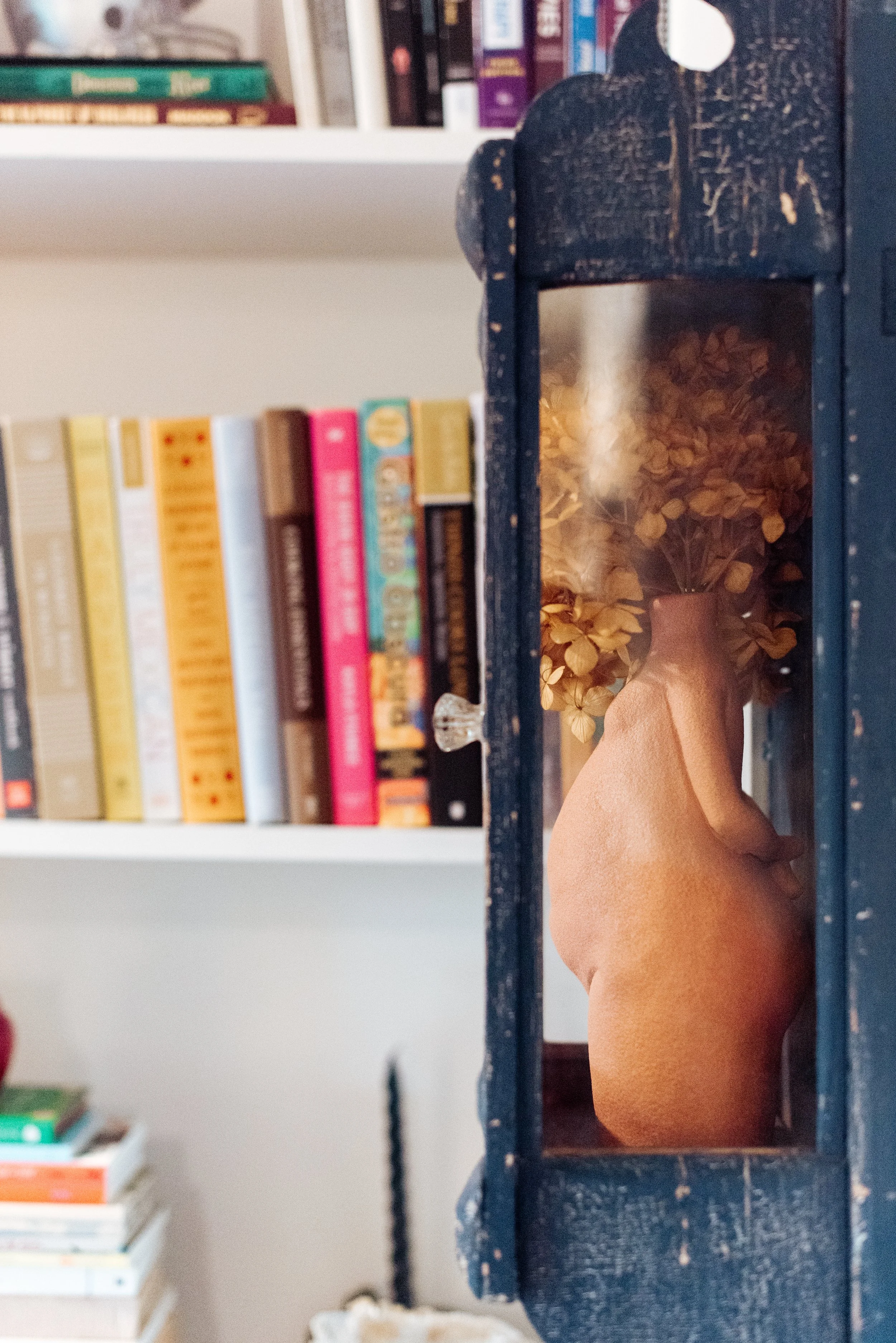 An orange ceramic vase with dried yellow flowers inside on a dark wooden shelf, with a bookshelf filled with books in the background.