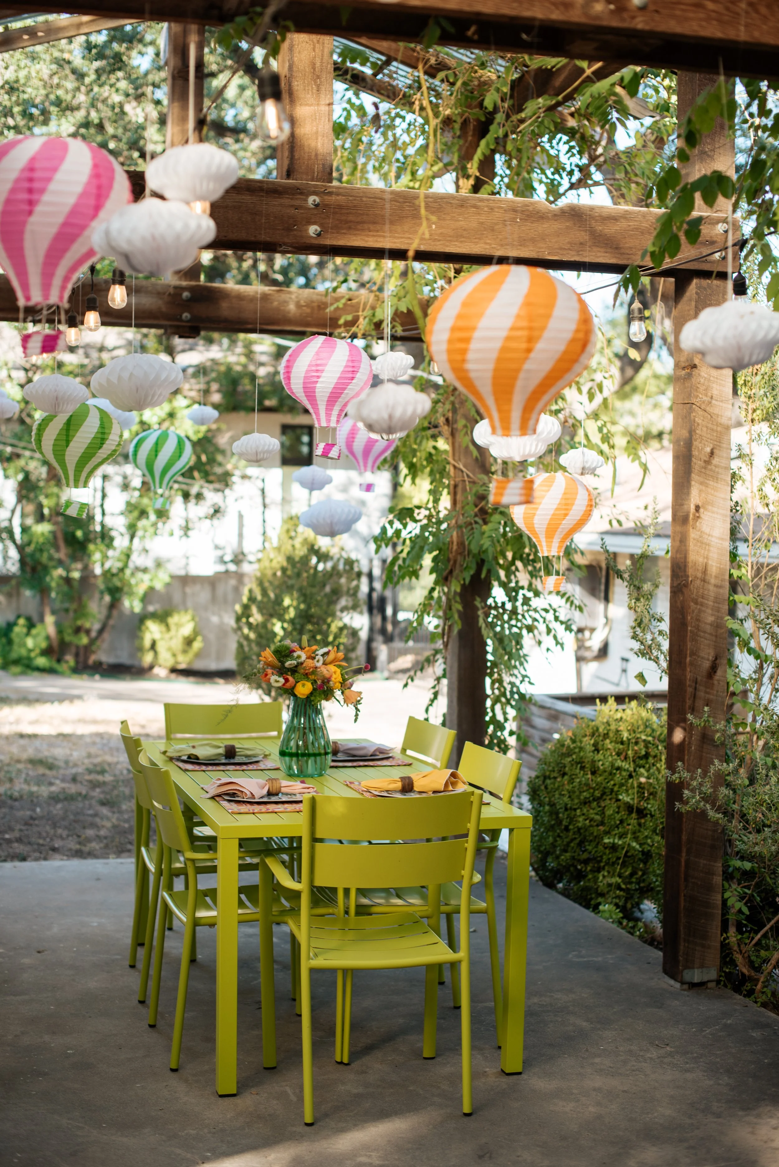 An outdoor dining area decorated with hanging paper lanterns and clouds in pink, orange, green, and white. A lime green table is set with napkins, cutlery, and a floral centerpiece in a green vase. The setting is surrounded by trees and plants, with 