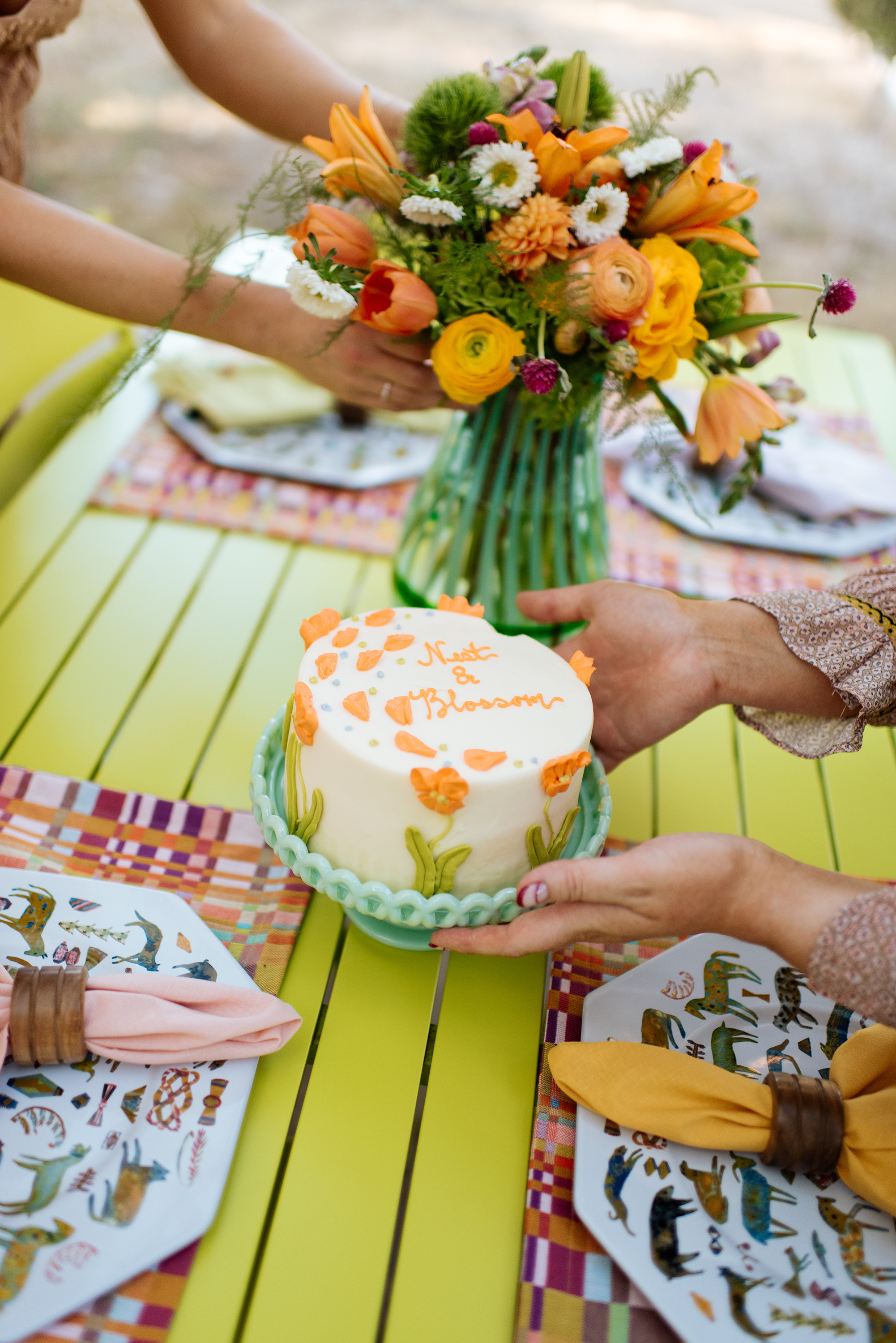 Person's hands holding a white cake decorated with orange flowers and leaves, with the words 'Next a Blossom' written on top. In the background, a large colorful flower arrangement in a green vase is on a yellow table with colorful placemats and decorative plates.