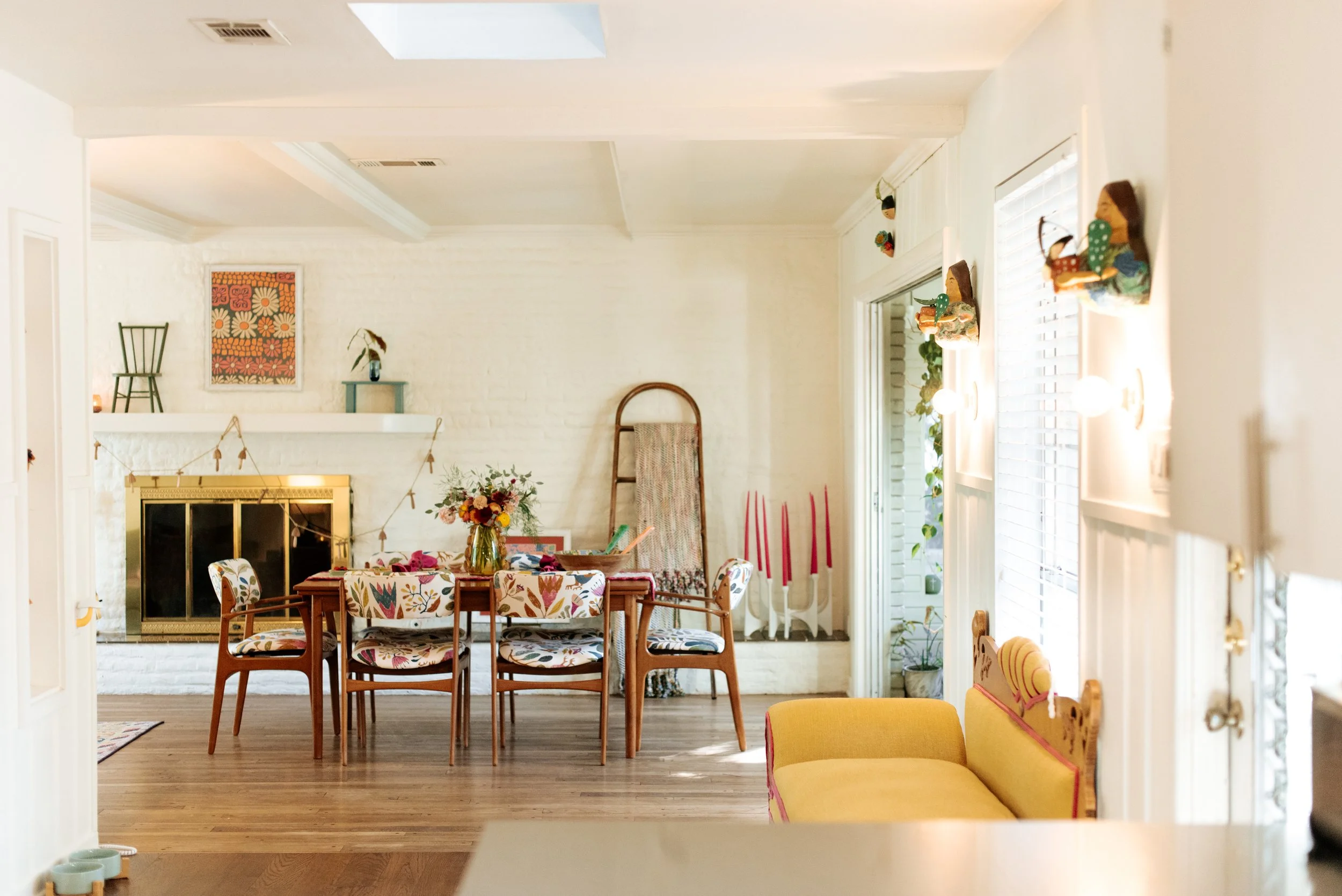 Bright and cheerful living room and dining area with a hardwood floor, white walls, and decorated with colorful artwork, flowers, candles, and vintage furniture including a yellow couch, a wooden dining table with floral chairs, and a cozy fireplace.