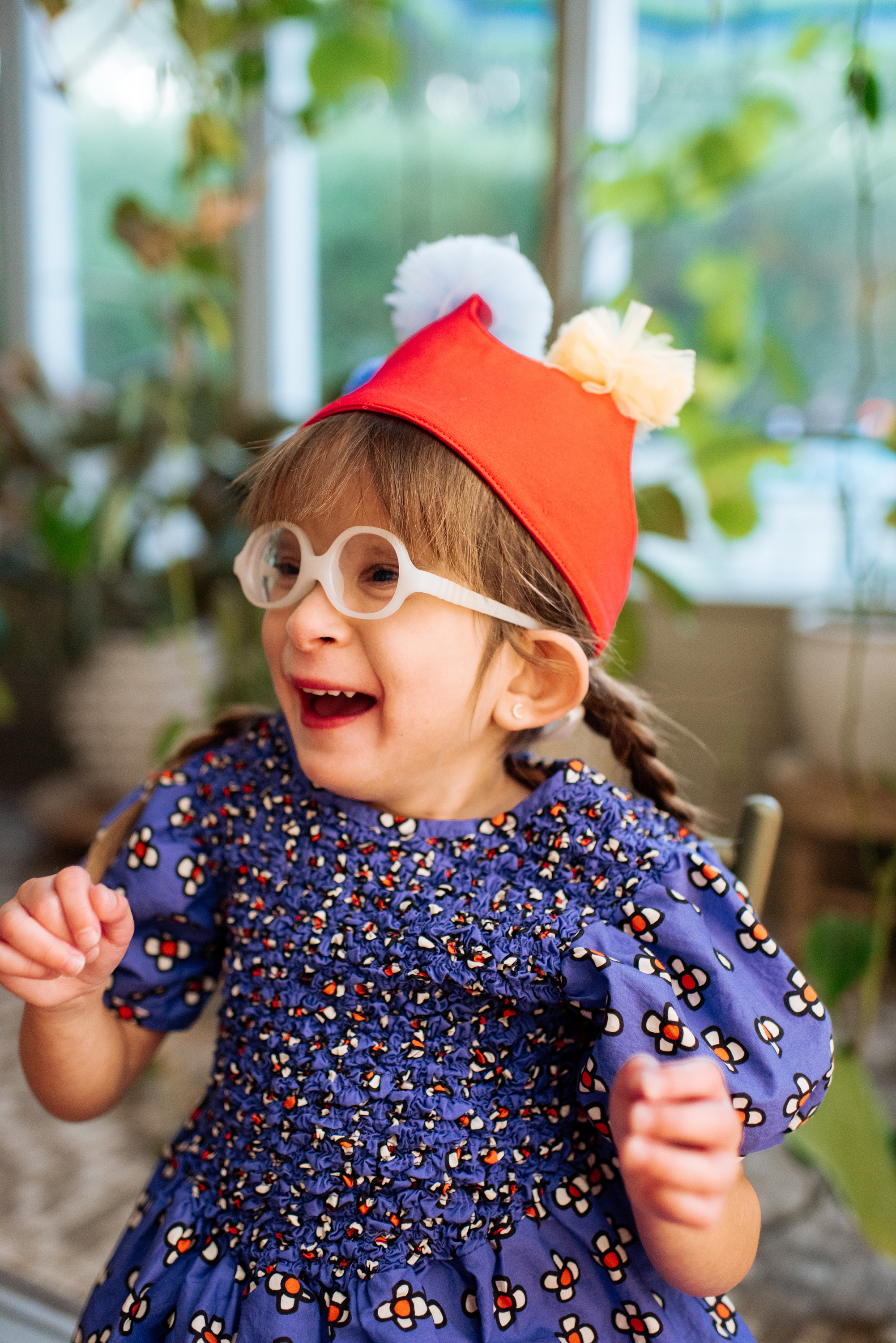 A happy young girl with glasses, wearing a colorful dress and a clown hat, laughing indoors near plants.