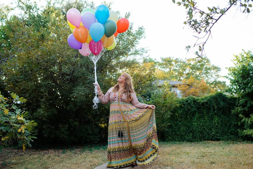 A woman holding a bunch of colorful balloons standing in a backyard with green trees and bushes.