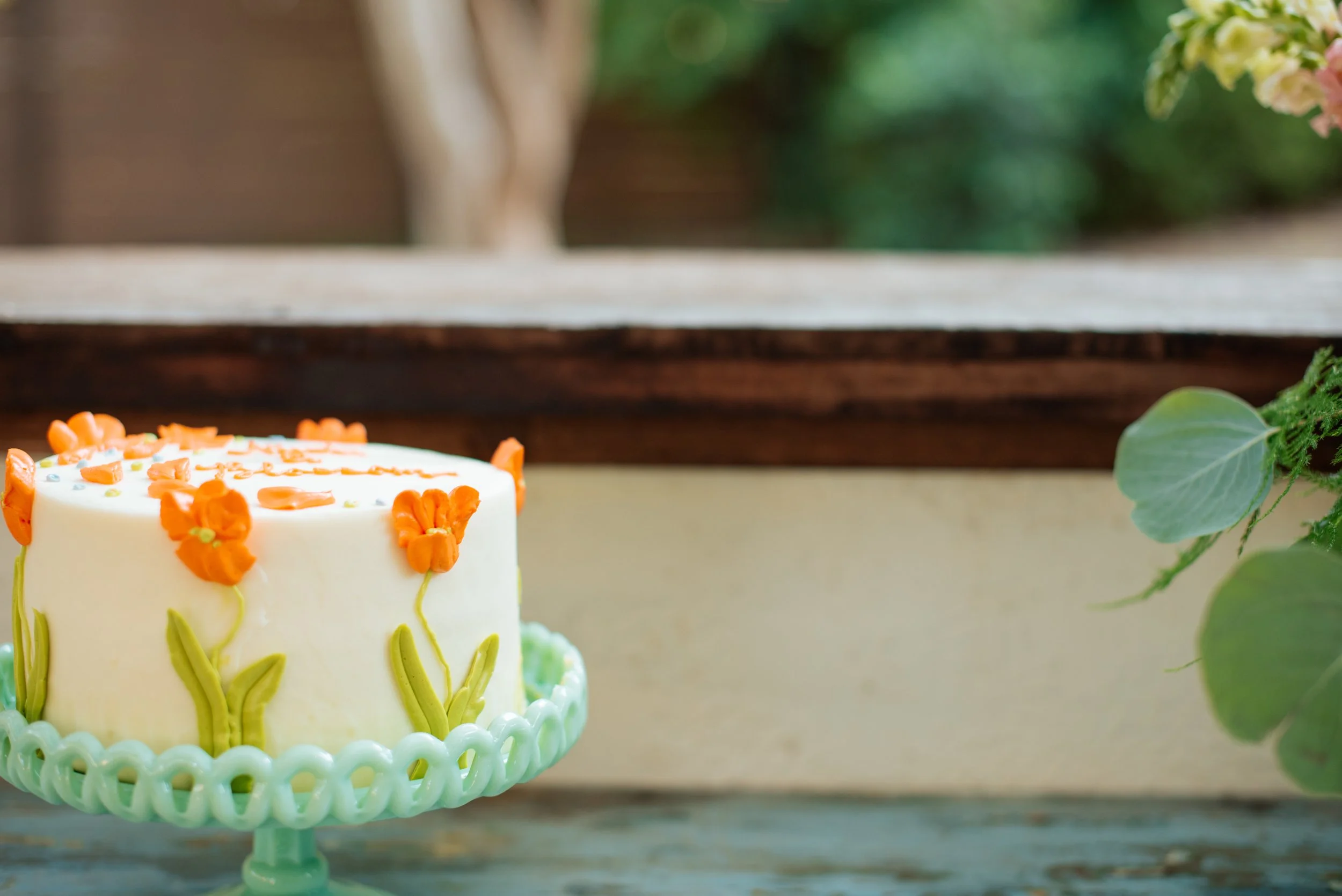 A white birthday cake decorated with orange flowers and green leaves on a mint green cake stand, placed outdoors.