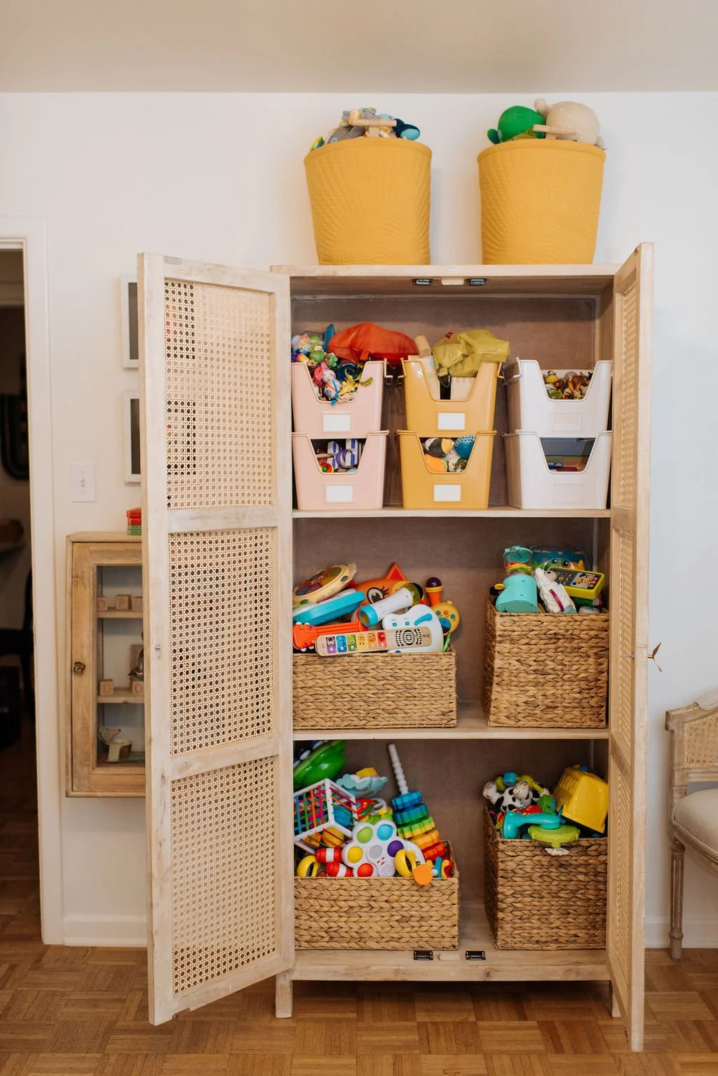 A wooden toy storage cabinet filled with various toys, including bins of colorful plastic toys, stuffed animals, and play sets, with two large yellow woven baskets on top.