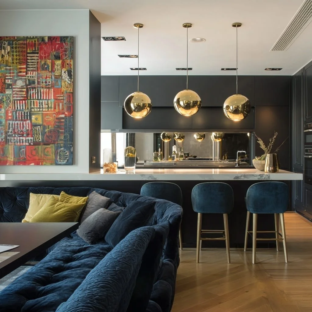 Vista del interior de una sala de estar que conecta con la cocina, con un sofá azul y cojines amarillos y grises, arte abstracto en la pared, barra de cocina con tres sillas de bar azul y lámparas colgantes doradas.
