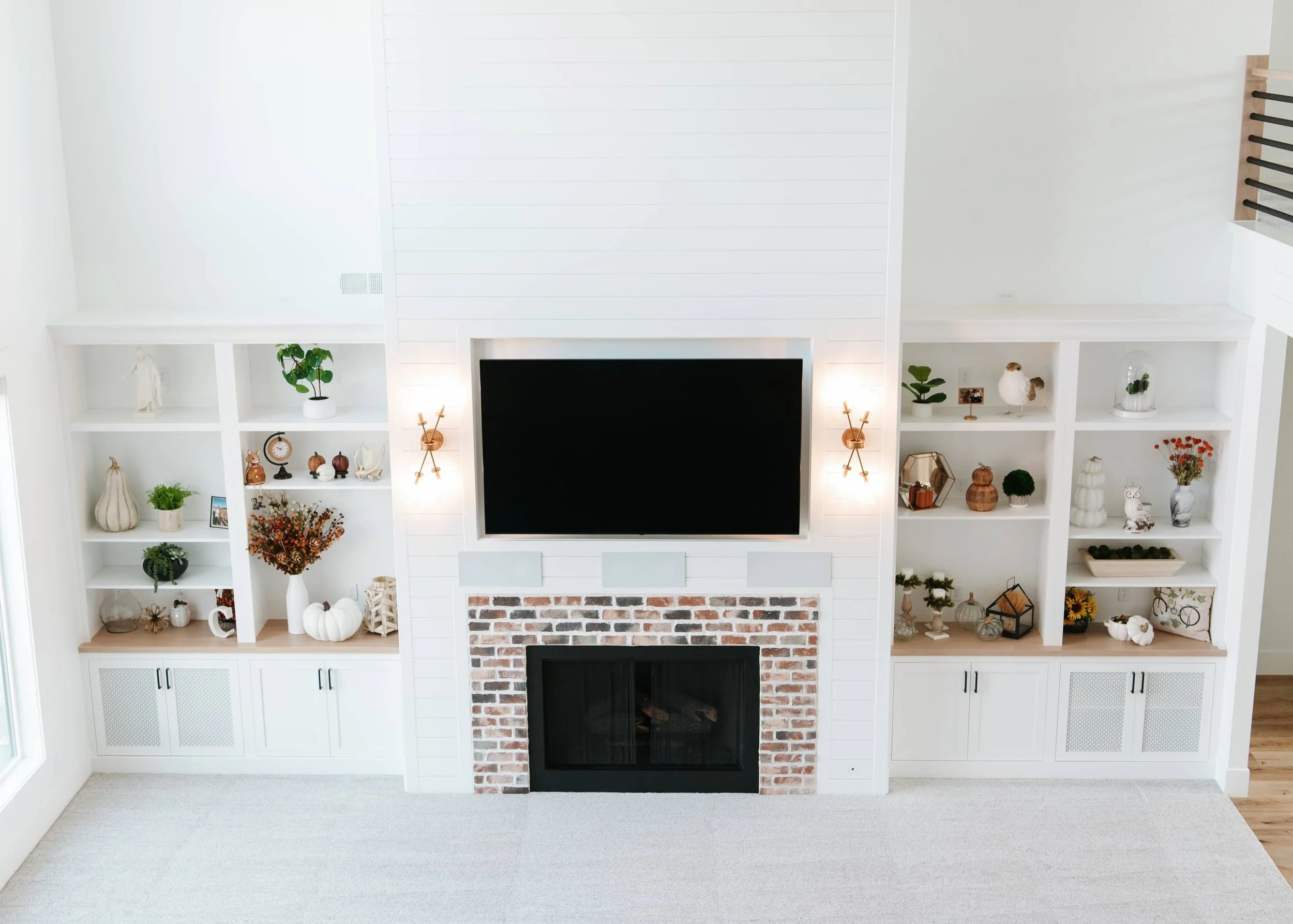 large white fireplace wall. The centerpiece is a brick-surround fireplace with a black insert, topped by a recessed flat-screen TV flanked by two glowing gold sconces. To the left and right are tall white built-in bookshelves filled with curated autu