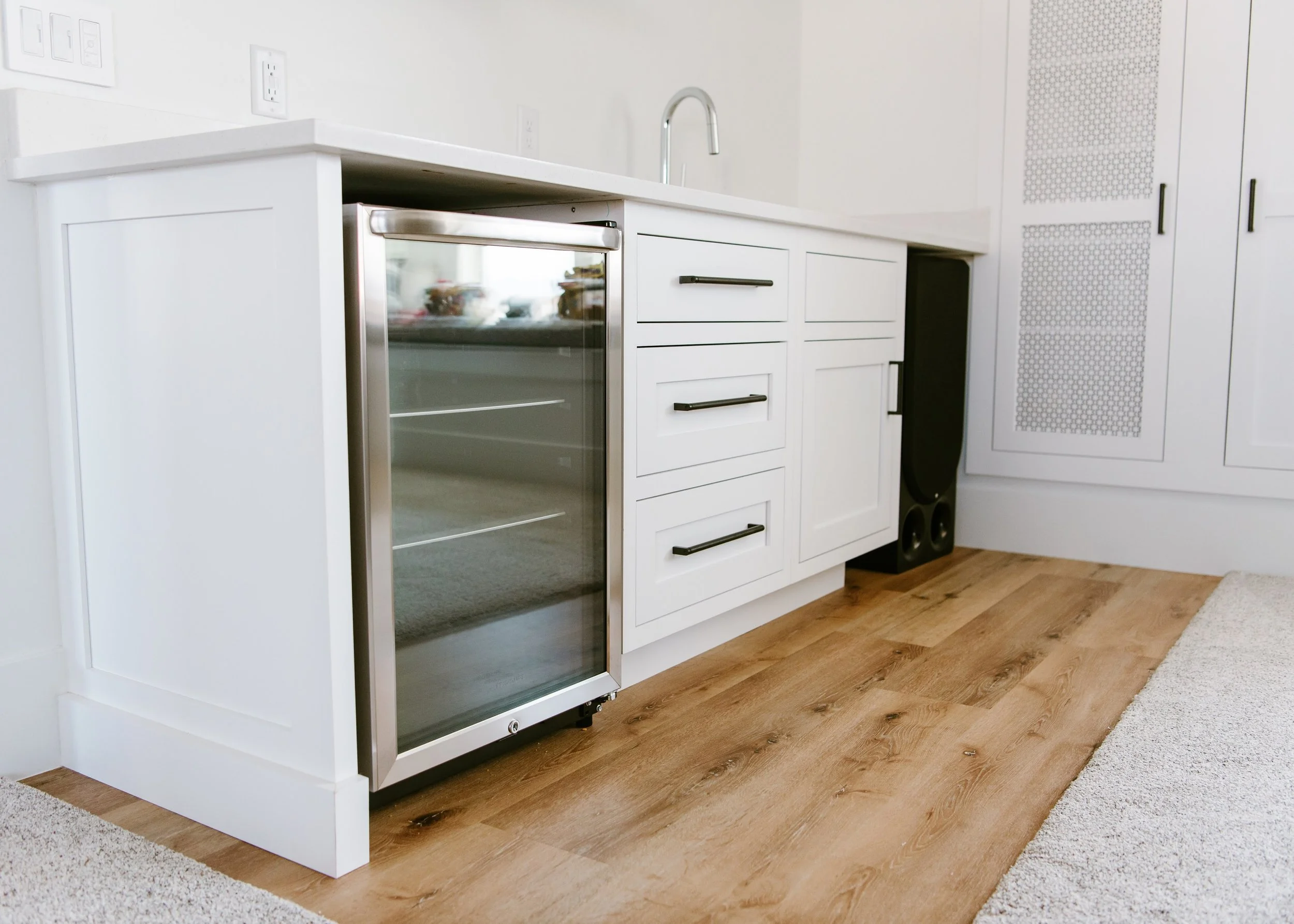 A modern white bar featuring shaker-style cabinetry with matte black handles and a white quartz countertop. A stainless steel under-counter beverage refrigerator with a glass door is built into the left side. The scene includes a chrome faucet, light