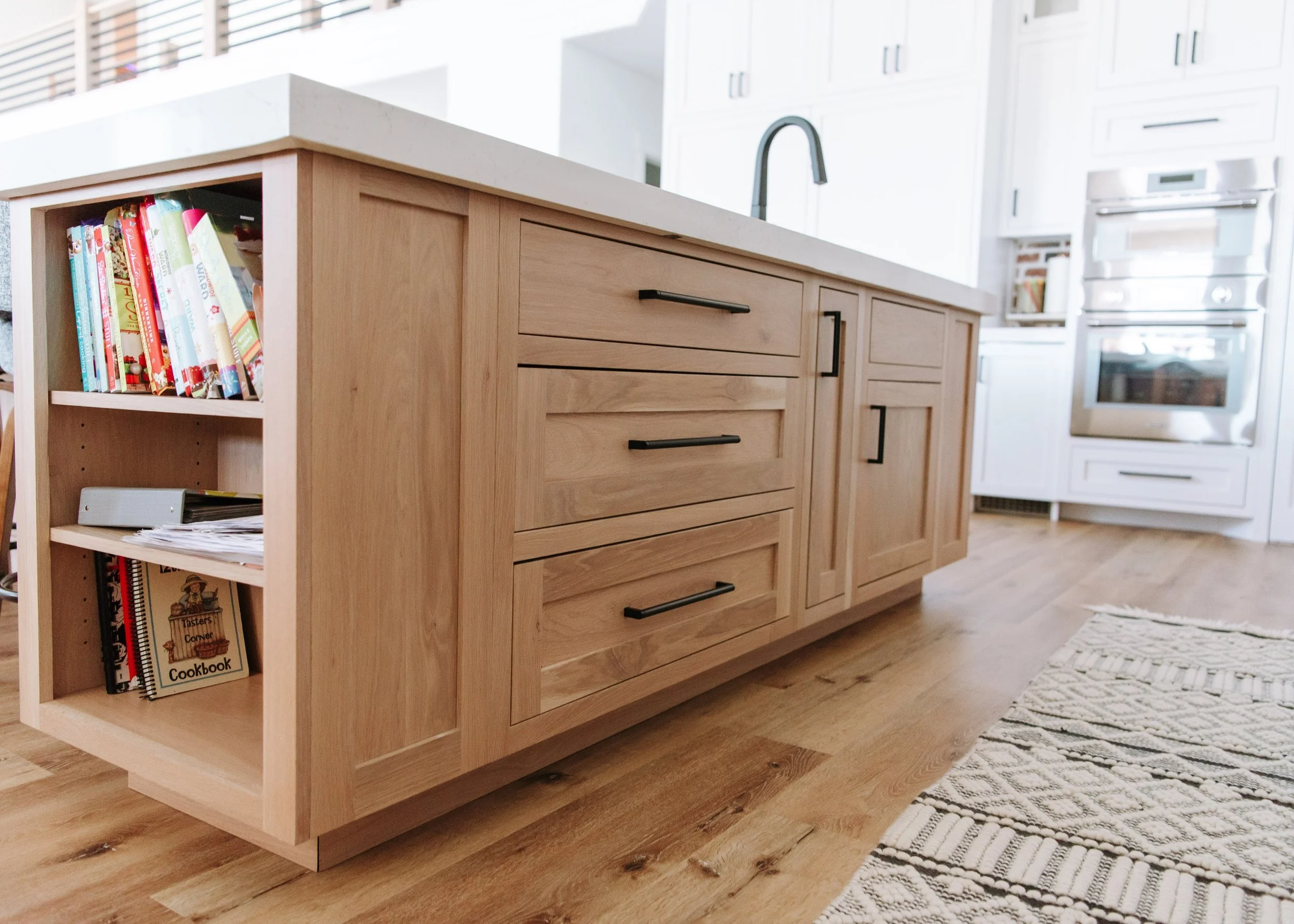 A close-up, low-angle shot of a light oak kitchen island featuring shaker-style drawers with matte black handles. The end of the island includes built-in open shelving filled with colorful cookbooks. The island is topped with a thick white quartz cou