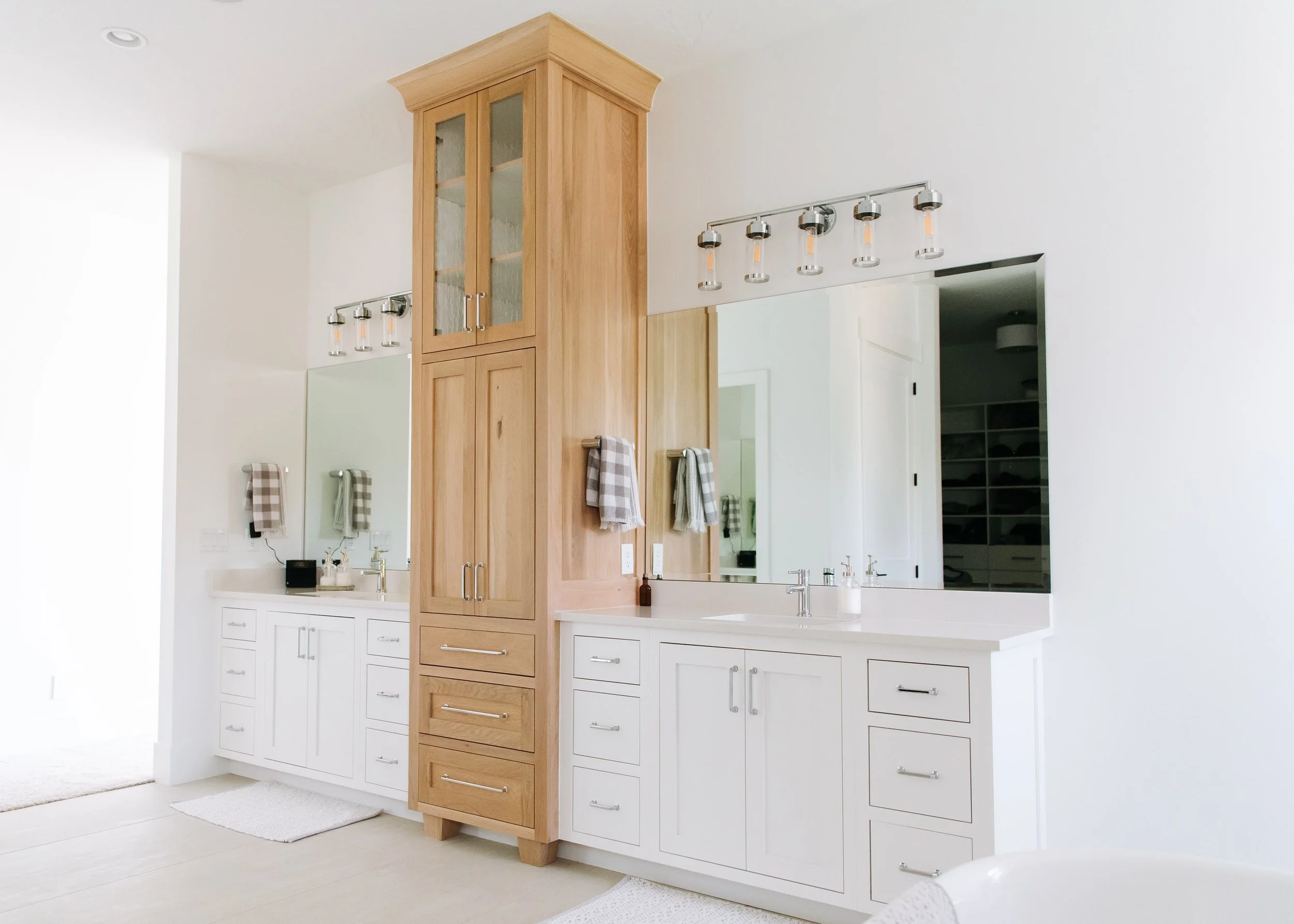 A bright, spacious primary bathroom featuring a double vanity with white cabinetry and a tall, central oak linen tower. The setup includes two separate sinks with large frameless mirrors and industrial-style chrome light fixtures above each. The desi
