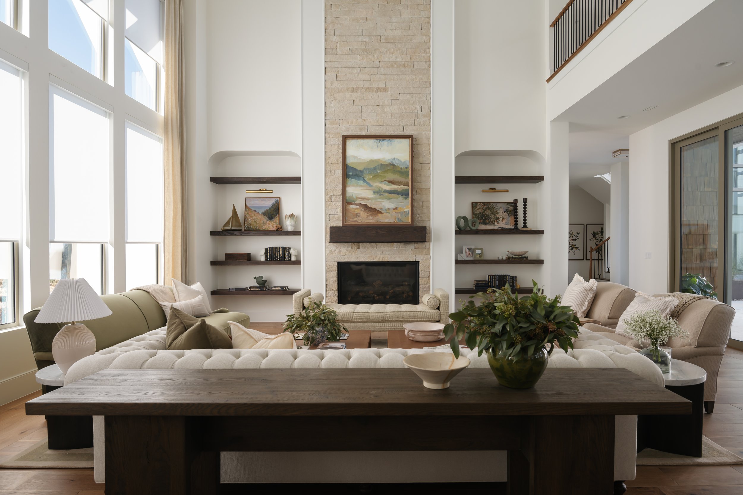 Bright living room with large windows, a fireplace, and built-in shelves filled with books and decor. Neutral-colored sofas with cushions and a wooden coffee table with plants and bowls. Modern, cozy, and stylish interior.