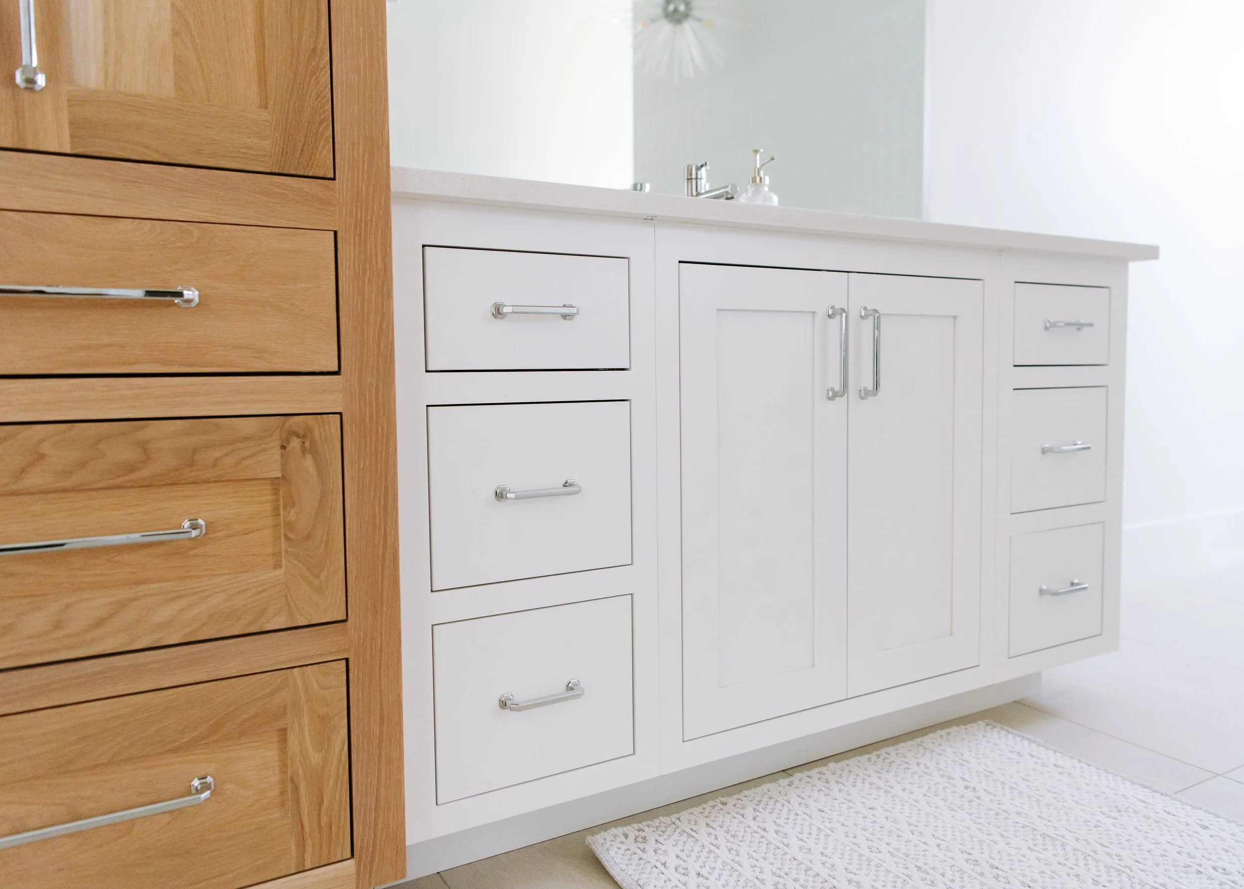 A detailed close-up of a bathroom vanity showing the contrast between natural oak drawers on the left and white shaker-style cabinets on the right. Both sections feature polished chrome hardware. A white quartz countertop and a textured white bath ma