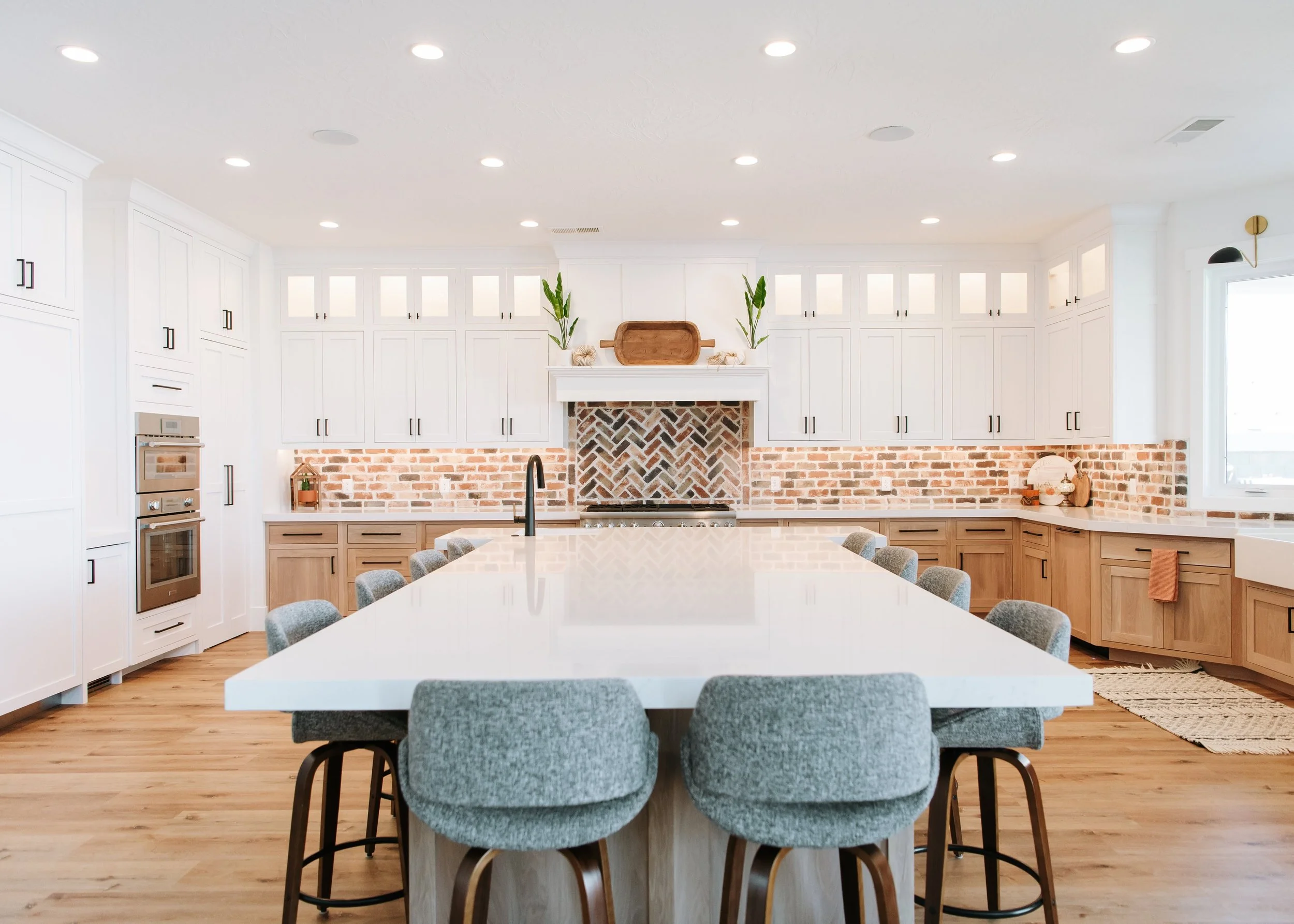 A wide, symmetrical shot of a spacious modern farmhouse kitchen. The design features white upper cabinetry, light wood lower cabinets, and a prominent brick backsplash with a herringbone pattern over the stove. A massive white-topped island with gray