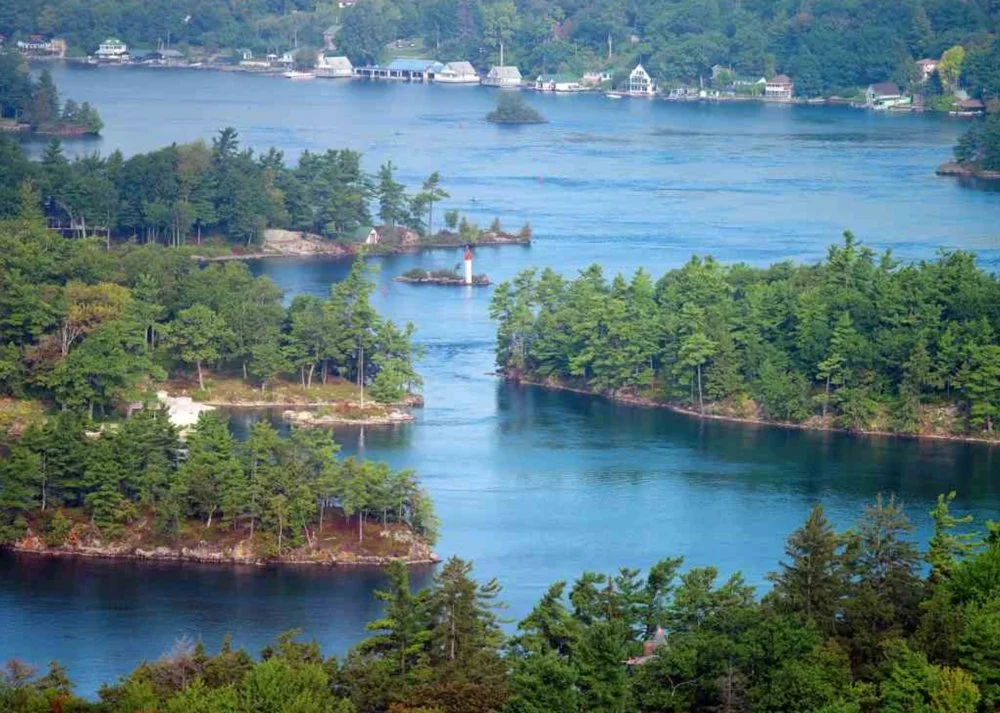 Landscape of a river with lush green trees on both sides, a lighthouse on a small island, and houses on the shoreline in the distance.