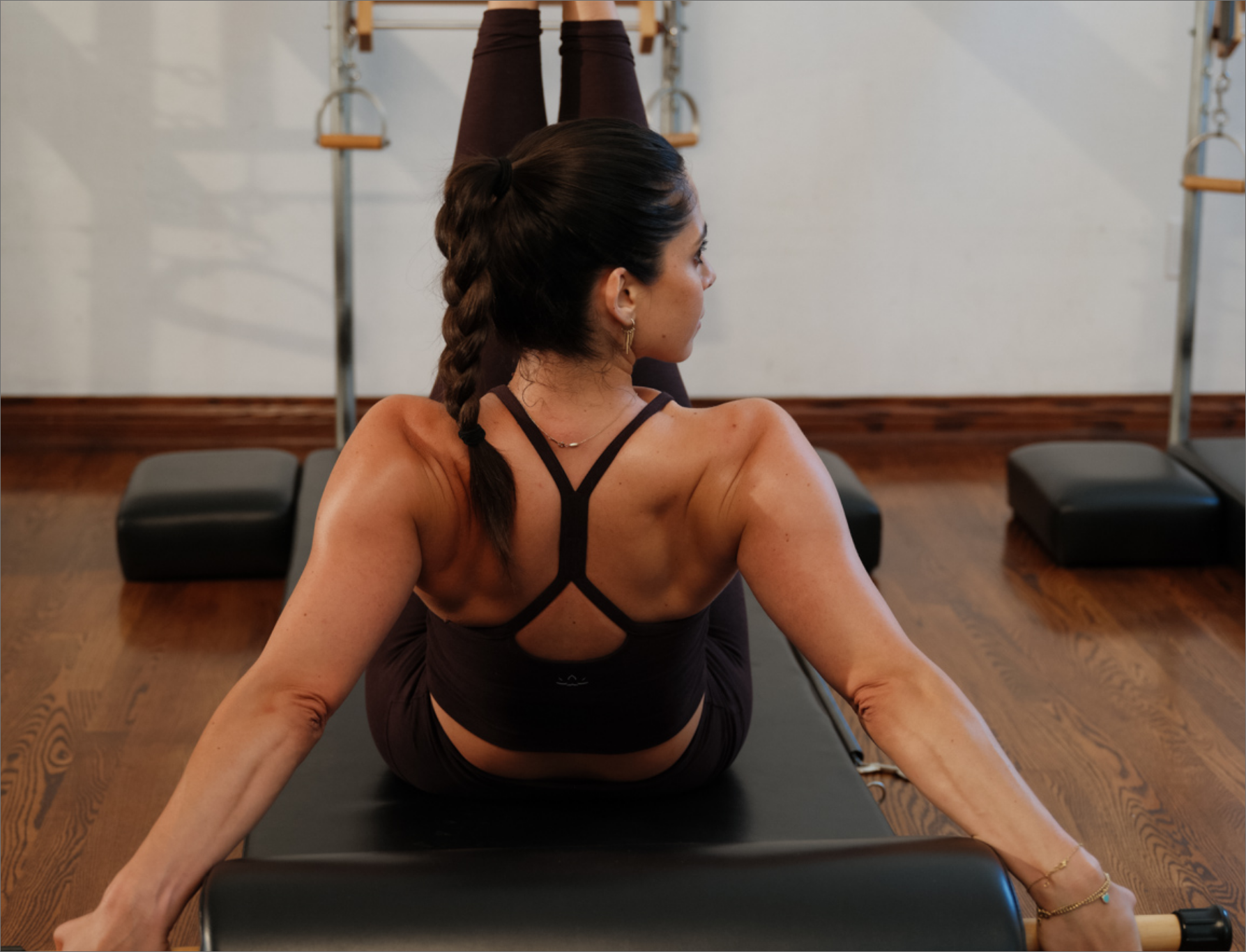 A woman practicing pilates on a black mat, sitting in a split pose with her back facing the camera, in a fitness studio with wooden flooring and wall-mounted equipment.