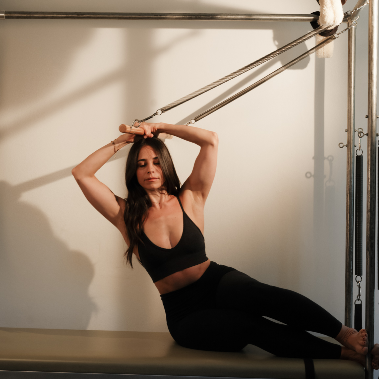 A woman in black workout attire stretching on a Pilates reformer machine, holding a wooden handle attached to springs, with her eyes closed and a focused expression.