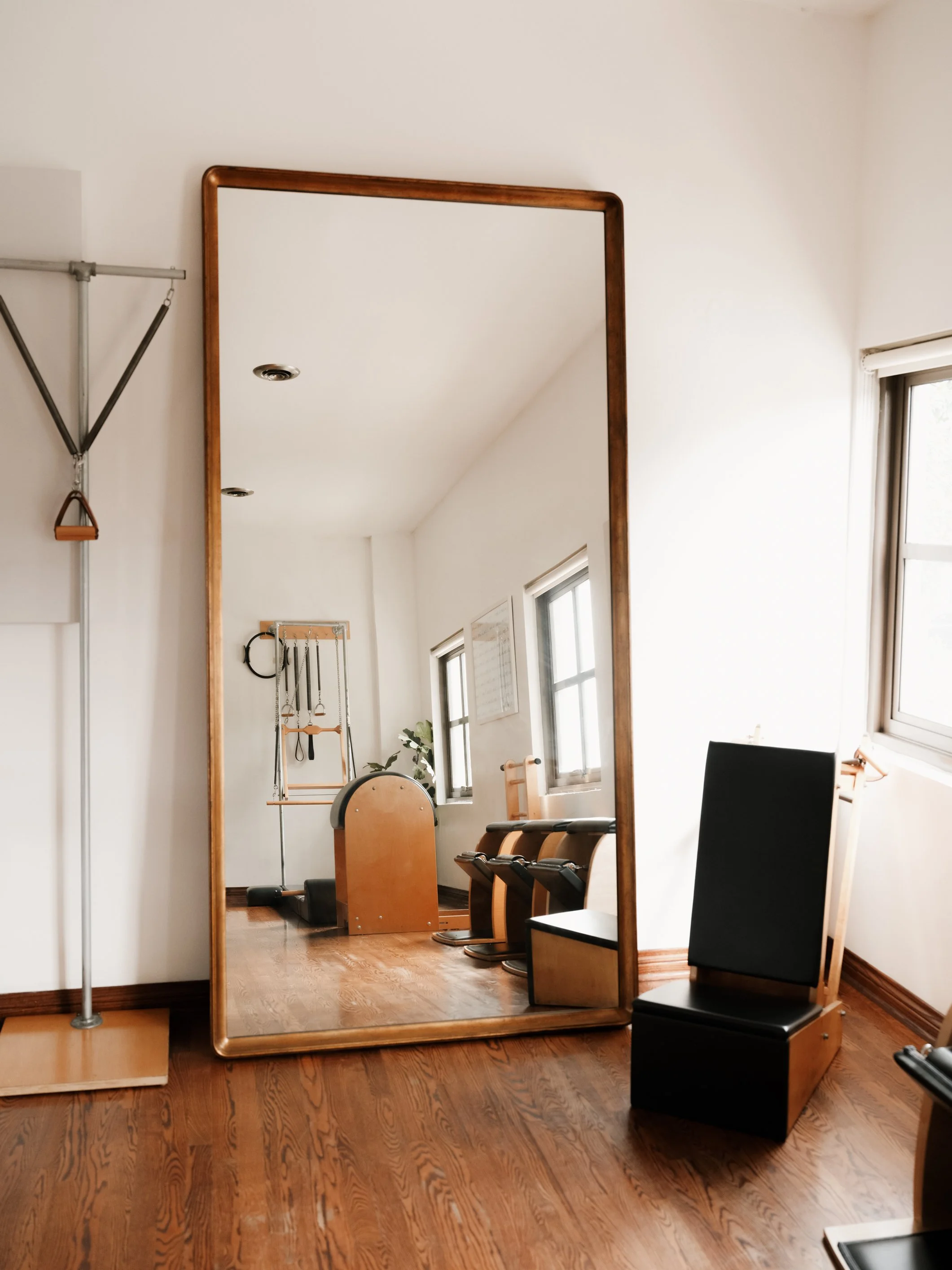 A large mirror in a room reflecting Pilates equipment and furniture, with wooden floors and windows.