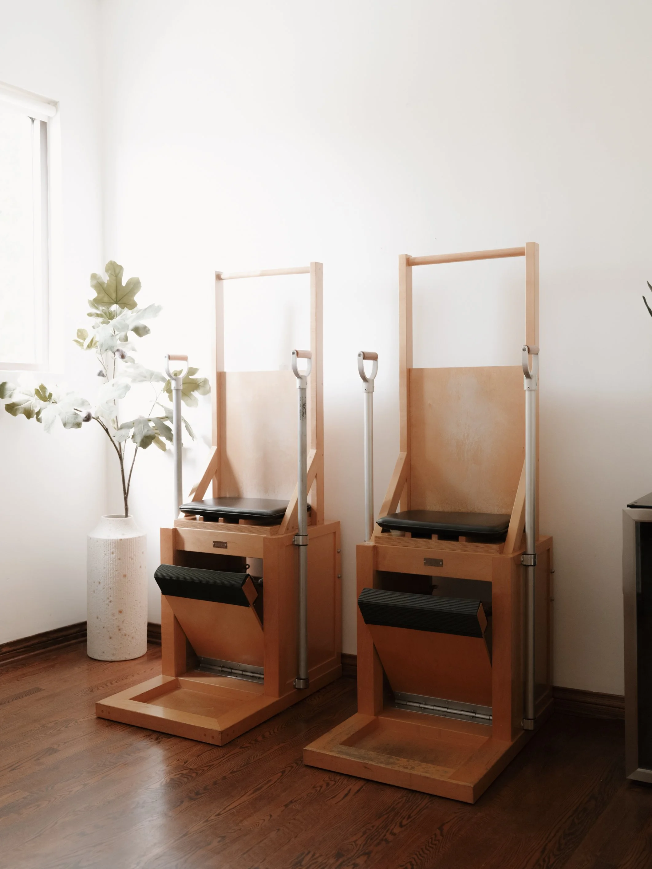 Two wooden Pilates chair machines in a bright room with hardwood floors, a potted plant, and a window.