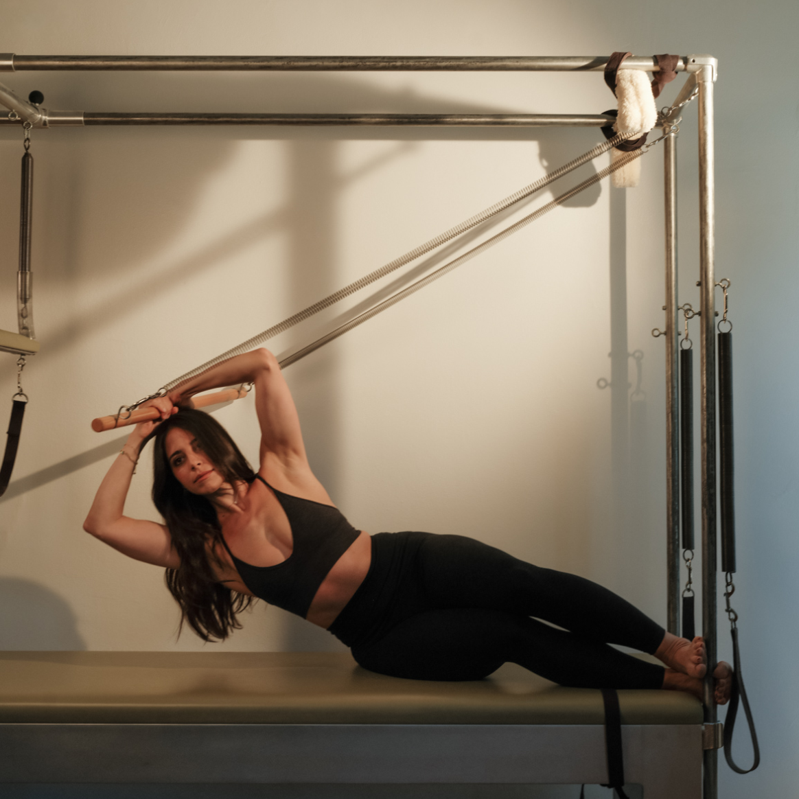 Woman with long dark hair in black workout clothes performing Pilates on a Cadillac machine.
