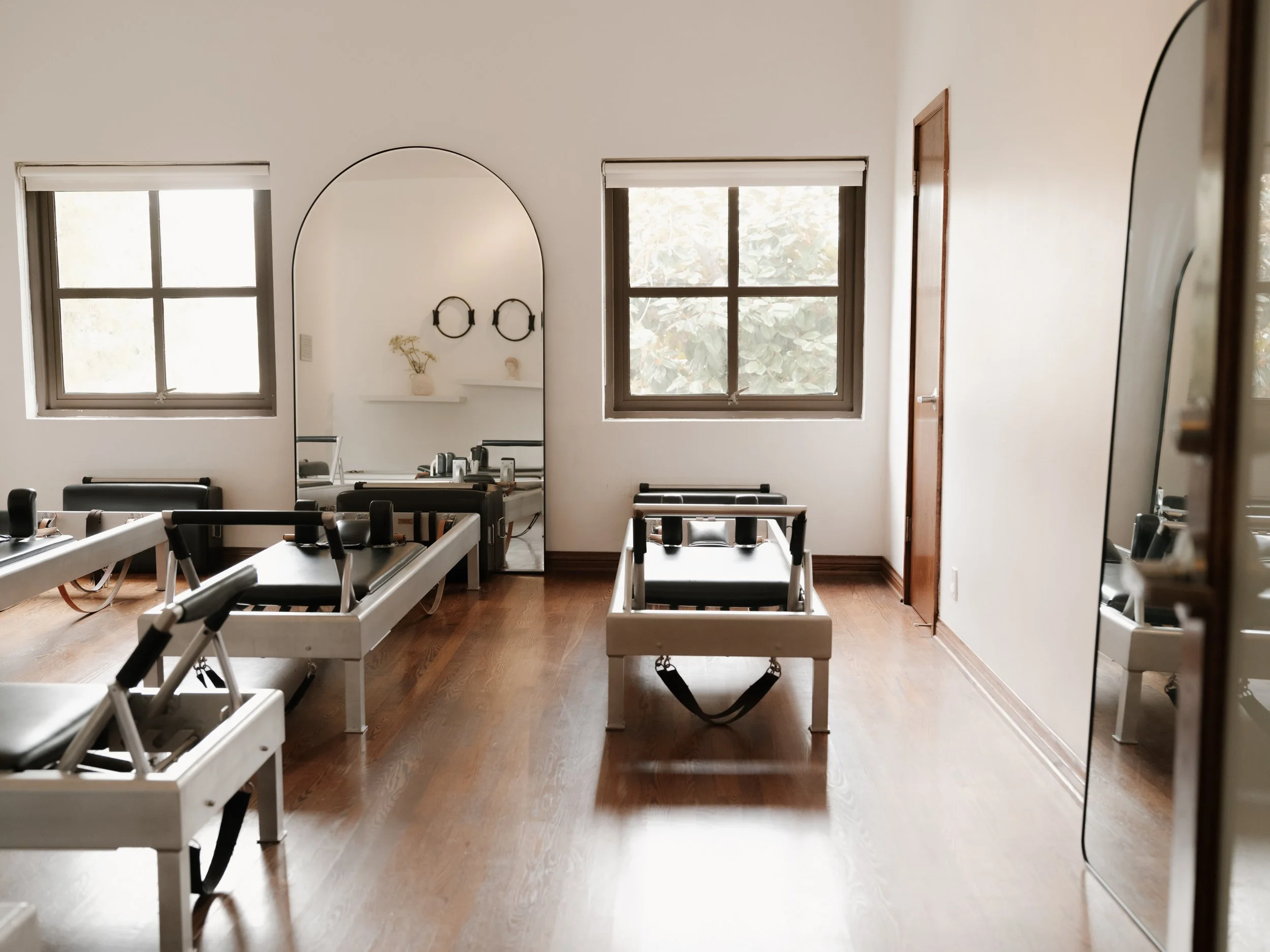 A bright room with wooden floors, two windows, a door, and a large mirror. There are four black and white Pilates reformer machines arranged in the room.