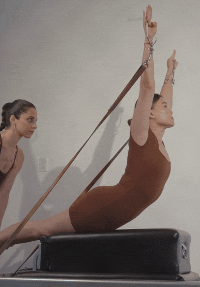 Two women performing exercises on a Pilates reformer machine. One woman is lying on her stomach, holding resistance straps, while the other woman stands nearby observing.
