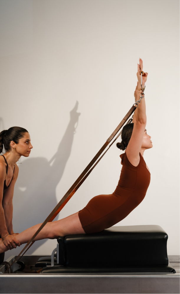 A woman performing an advanced Pilates exercise on a reformer machine, with assistance from an instructor.