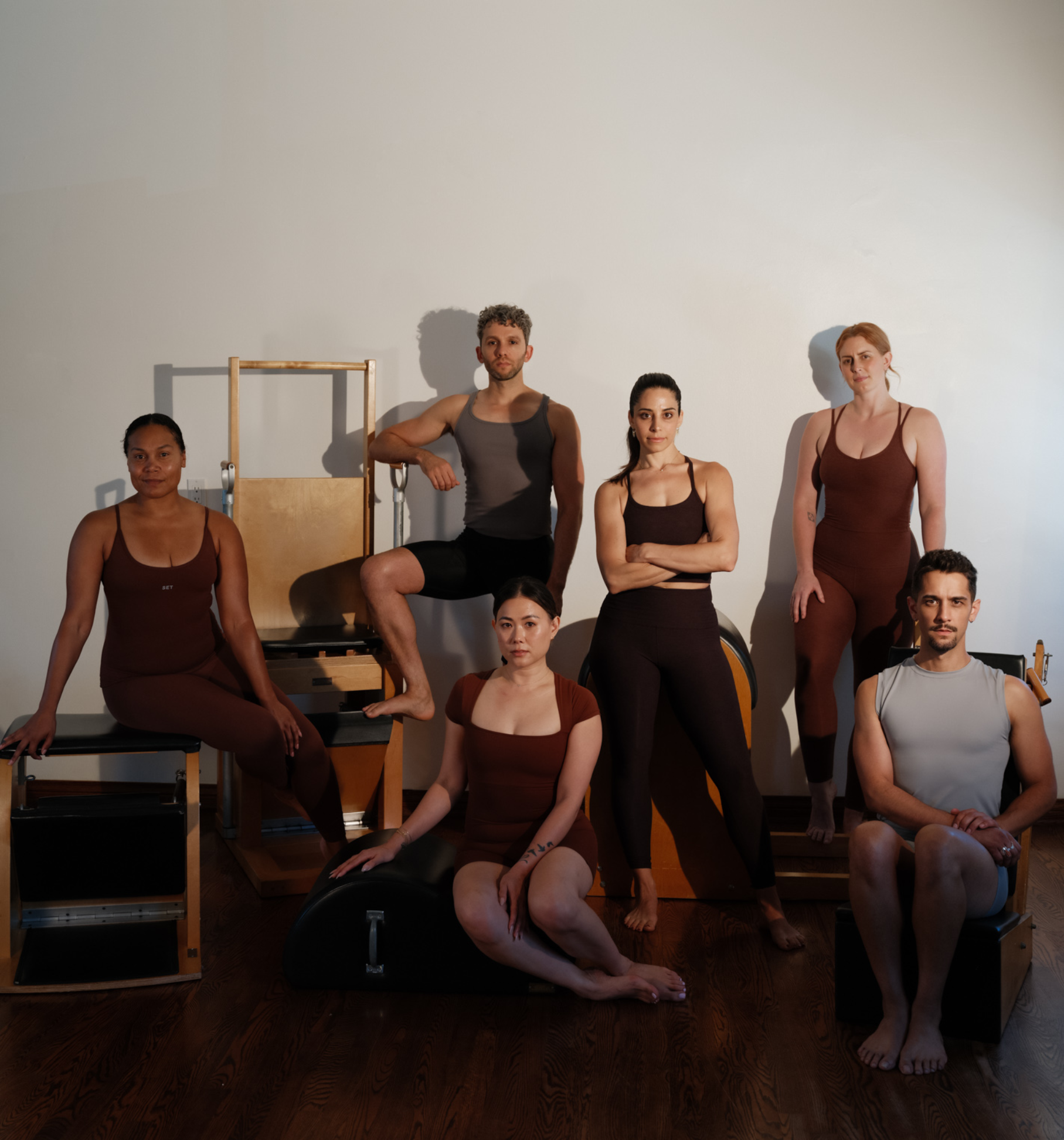 Group of seven diverse individuals in workout attire posing in a fitness studio with Pilates equipment.