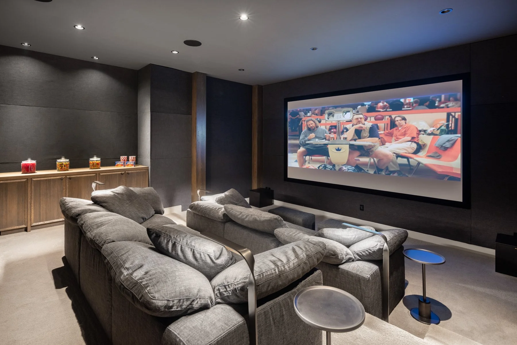 A home theater room with a large screen, gray sectional sofa, small side tables, and a cabinet with jars of candy along the back wall.