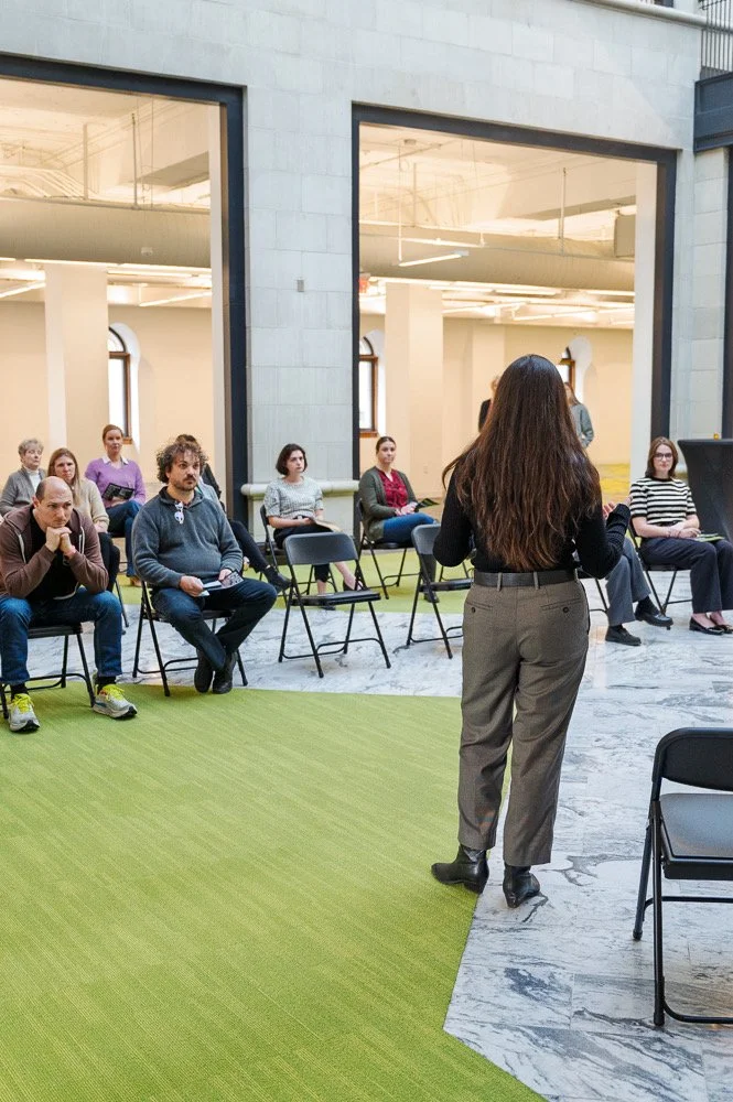 A woman with long brown hair, wearing gray pants and a black top, is speaking to a group of seated people in a spacious, well-lit auditorium with large windows and a mix of green carpet and marble flooring.