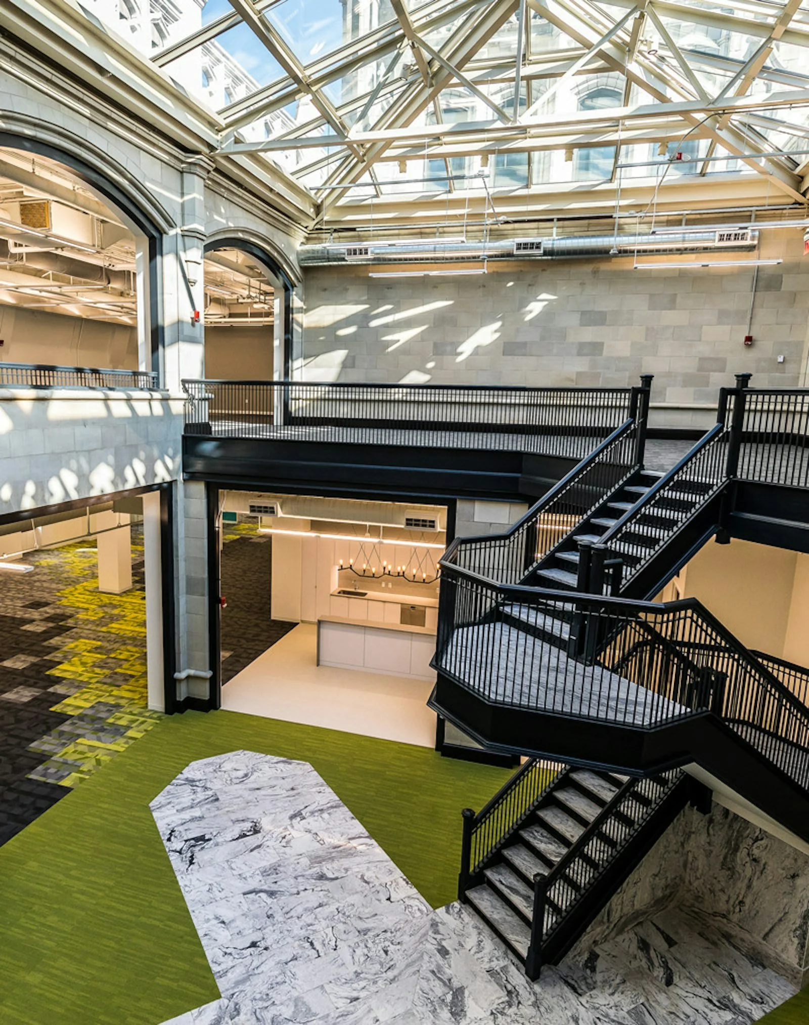 Interior of a modern building with a glass ceiling, stone walls, and black metal staircase. The floor features a pattern with green, black and white marble tiles.