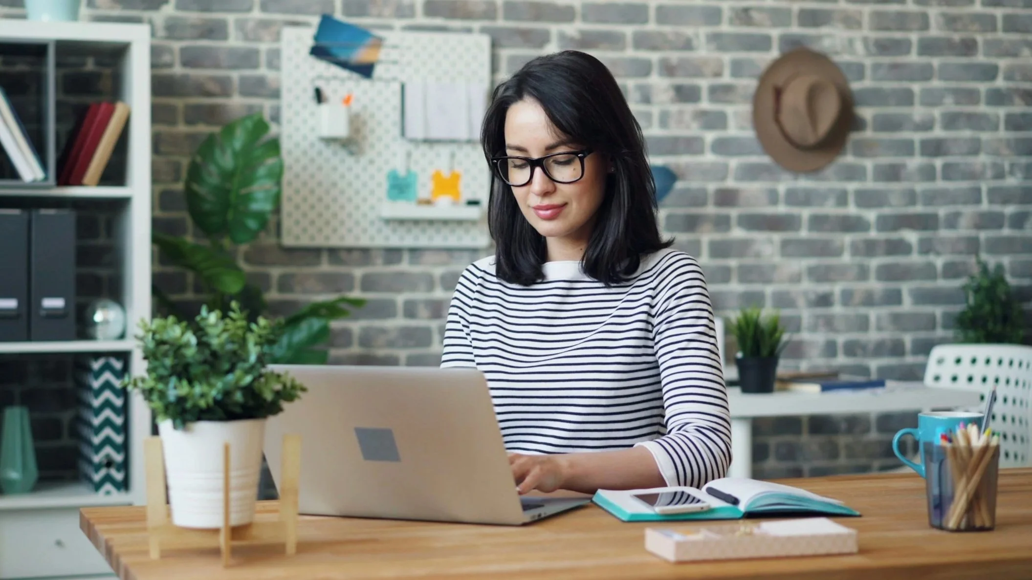 A woman with black hair and occupational glasses working on a laptop at a desk in a modern office with plants, books, and a brick wall background.
