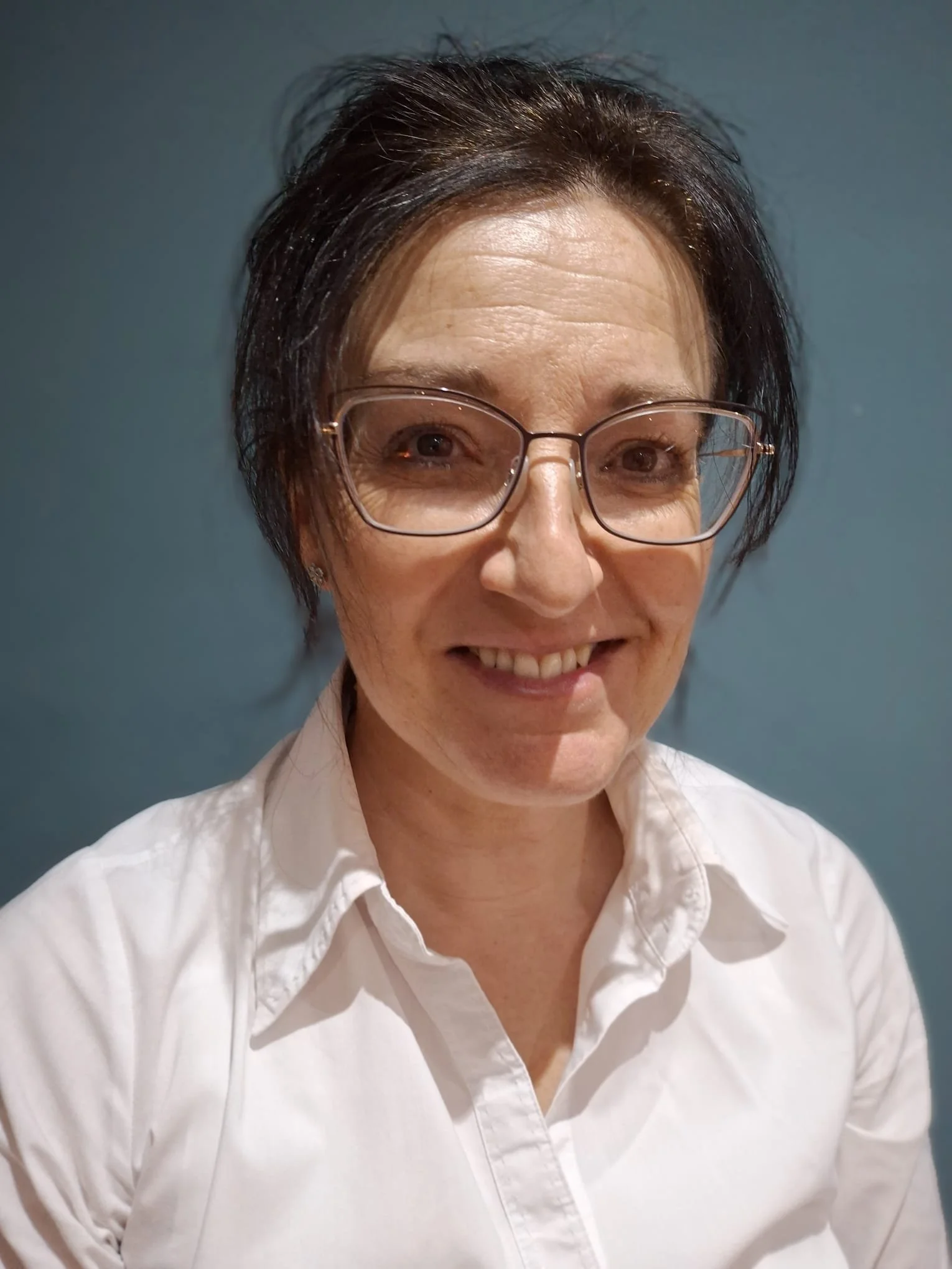 An optometrist with dark hair, wearing glasses and a white button-up shirt, smiling at the camera against a plain blue-grey background.