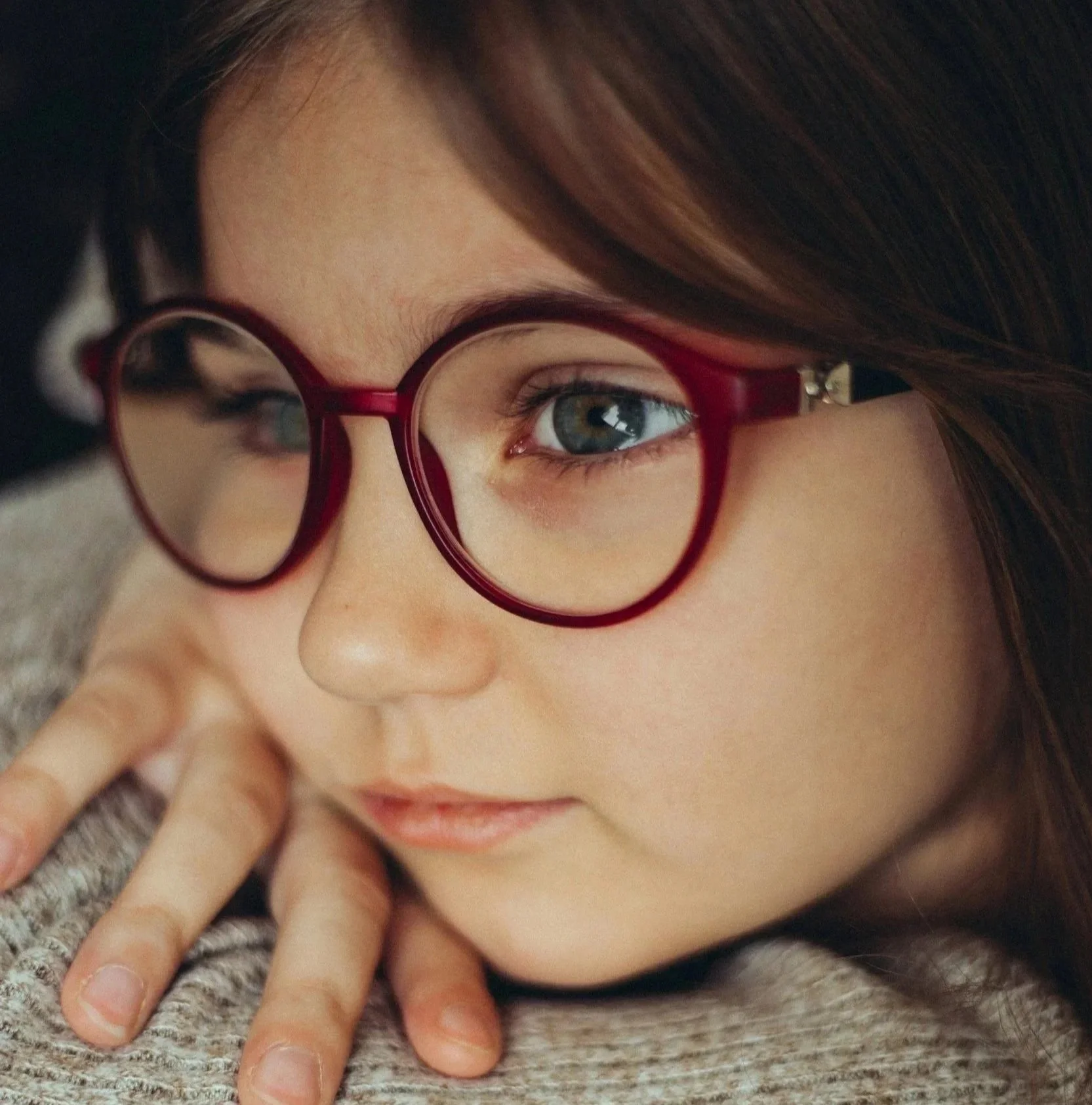 Close-up of a young girl with blue eyes and long brown hair, wearing red round glasses with myopia control lenses, laying on a textured surface.