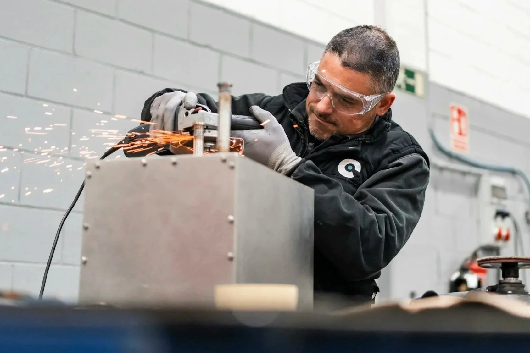 A man wearing safety goggles and gloves welding metal, sparks flying as he works on a metal box in an industrial setting.