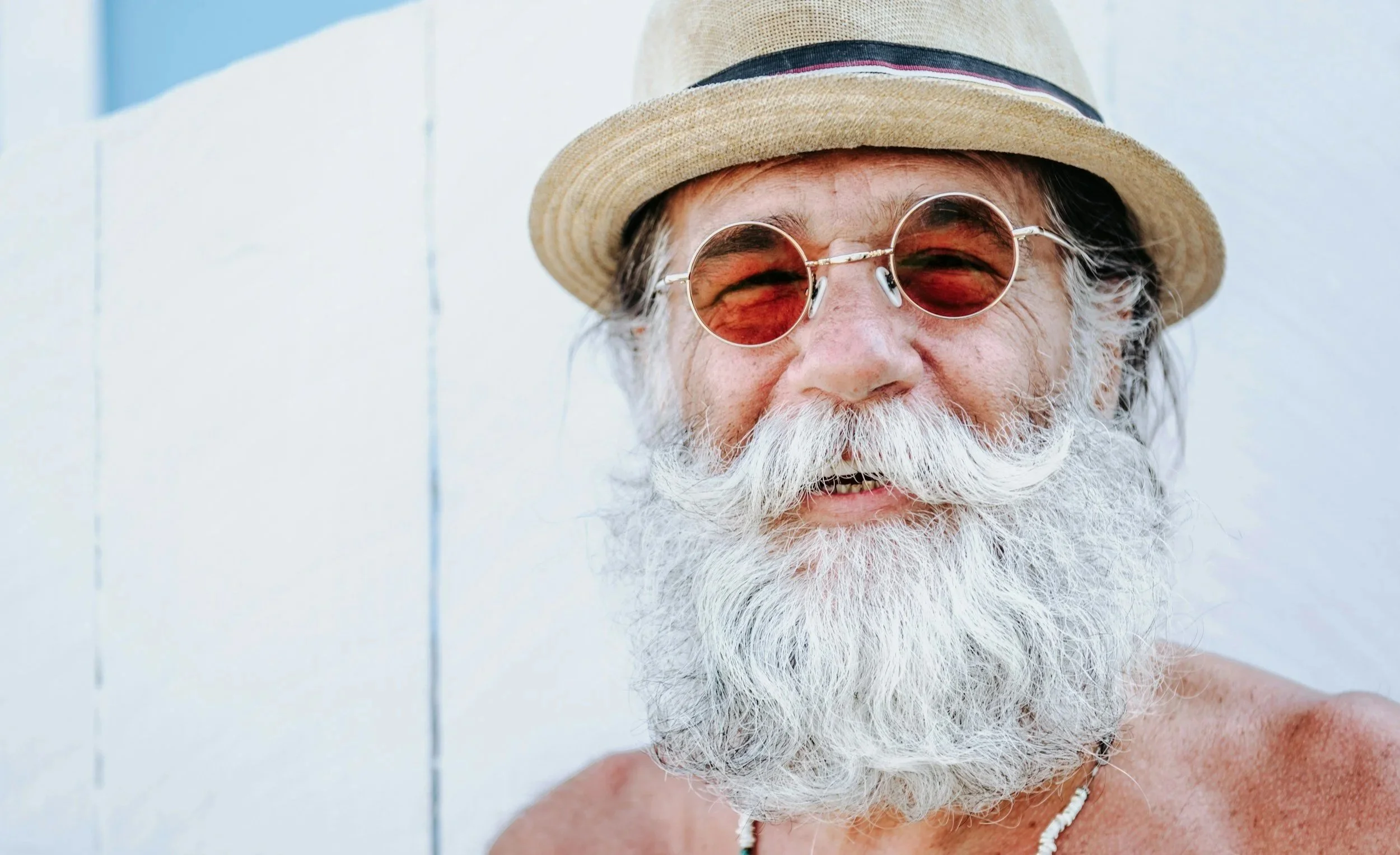A smiling elderly man with a long white beard, wearing sunglasses, a straw hat, and a necklace, standing against a white wall.