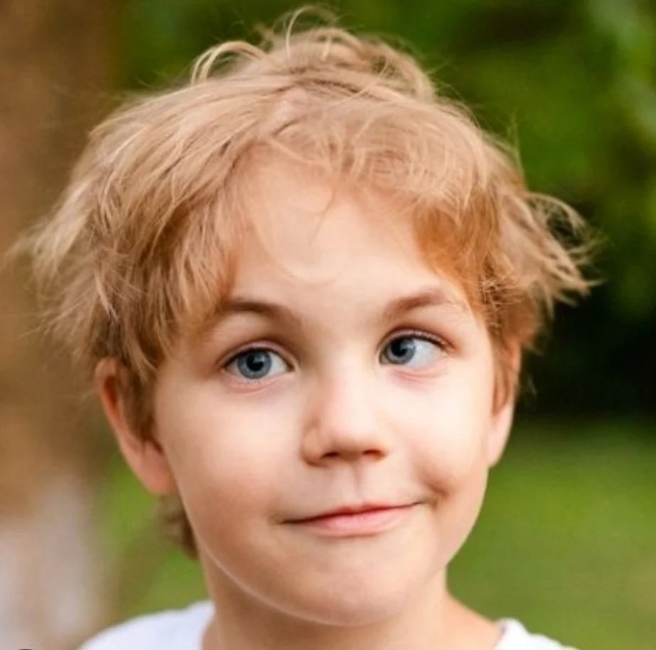 Close-up of a young boy with wavy blonde hair and blue eyes, outdoors with blurred green background.