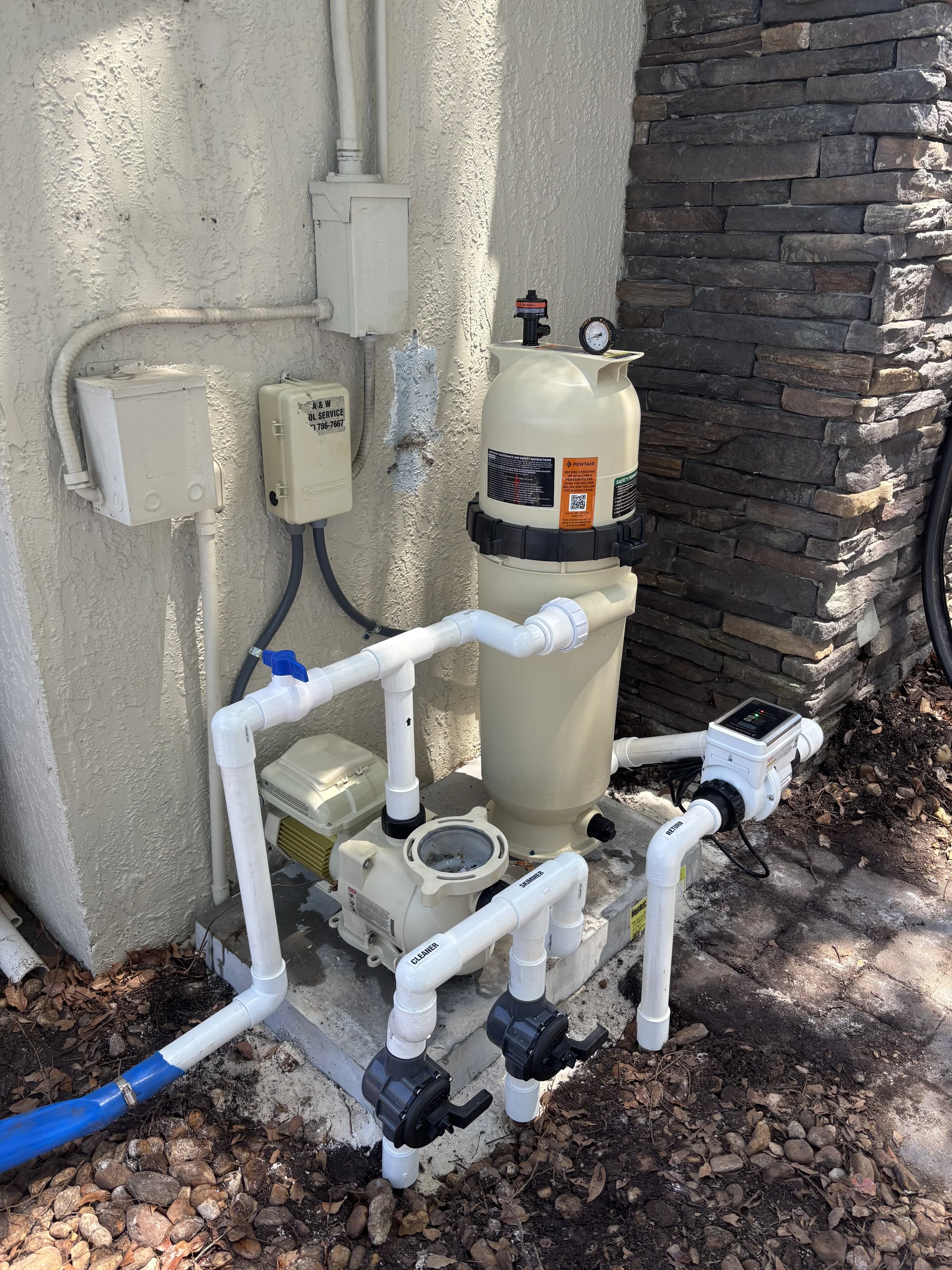 A backyard pool pump system with a filter, motor, pipes, valves, and an automatic controller installed outside next to a house wall and a brick pillar.