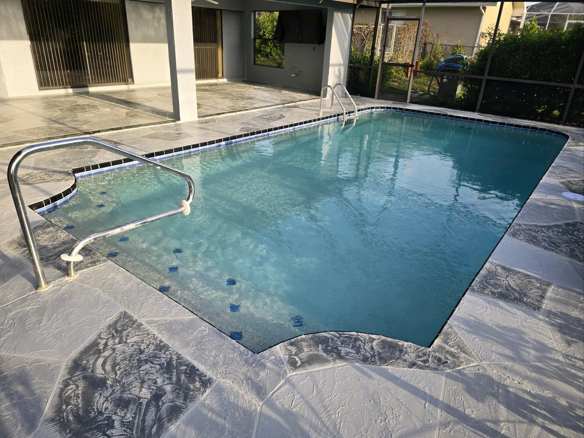 An outdoor swimming pool with metal handrails, surrounded by a textured stone deck, enclosed in a screened area with a house and garden in the background.