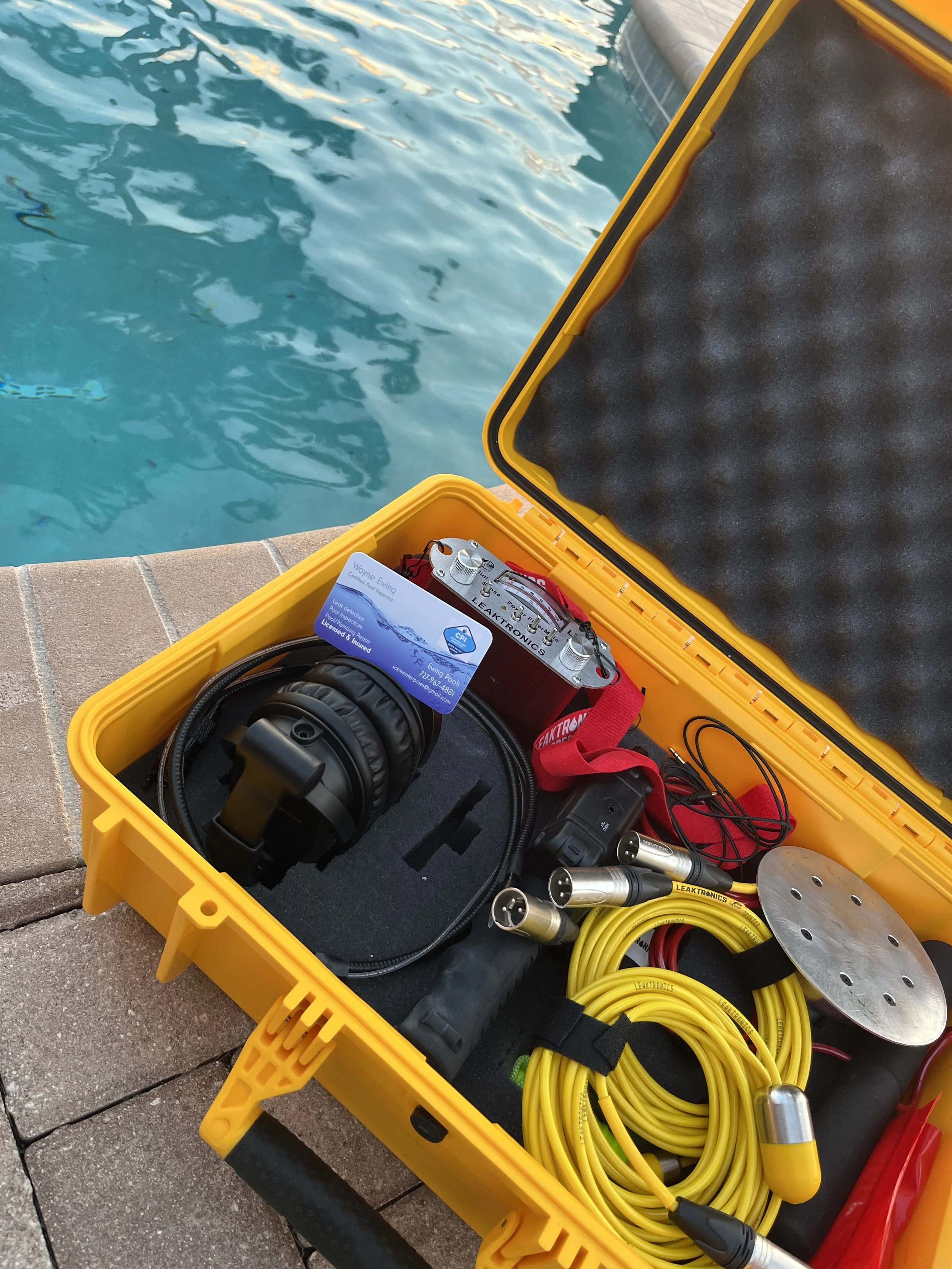 A yellow waterproof case containing electronic equipment like cables, a headlamp, and a metal disk, placed on a dock near a swimming pool.