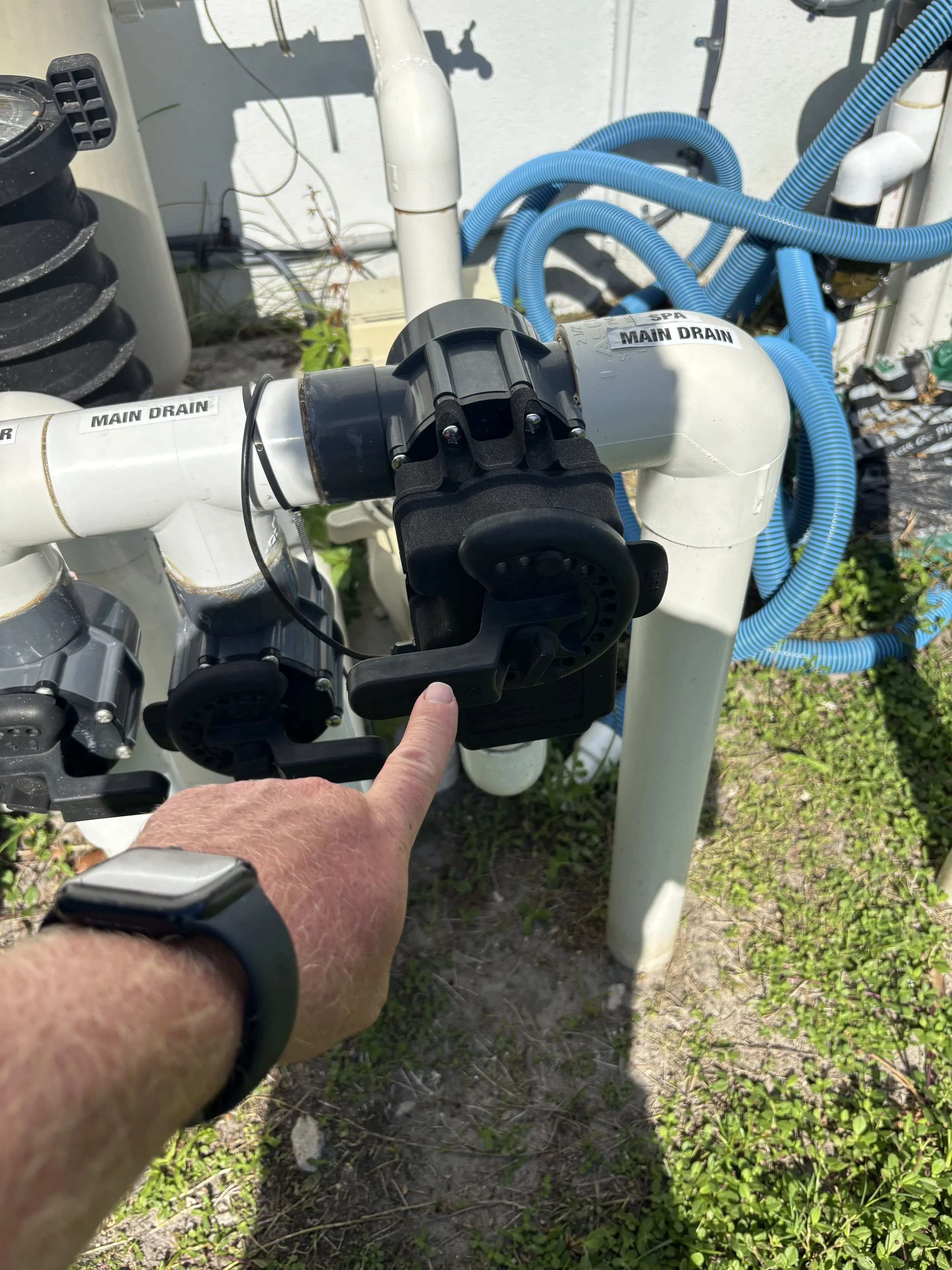 Close-up of a person pressing a switch on a large pool pump system with labeled pipes, surrounded by green grass and concrete, with blue flexible pipes attached to the pump.