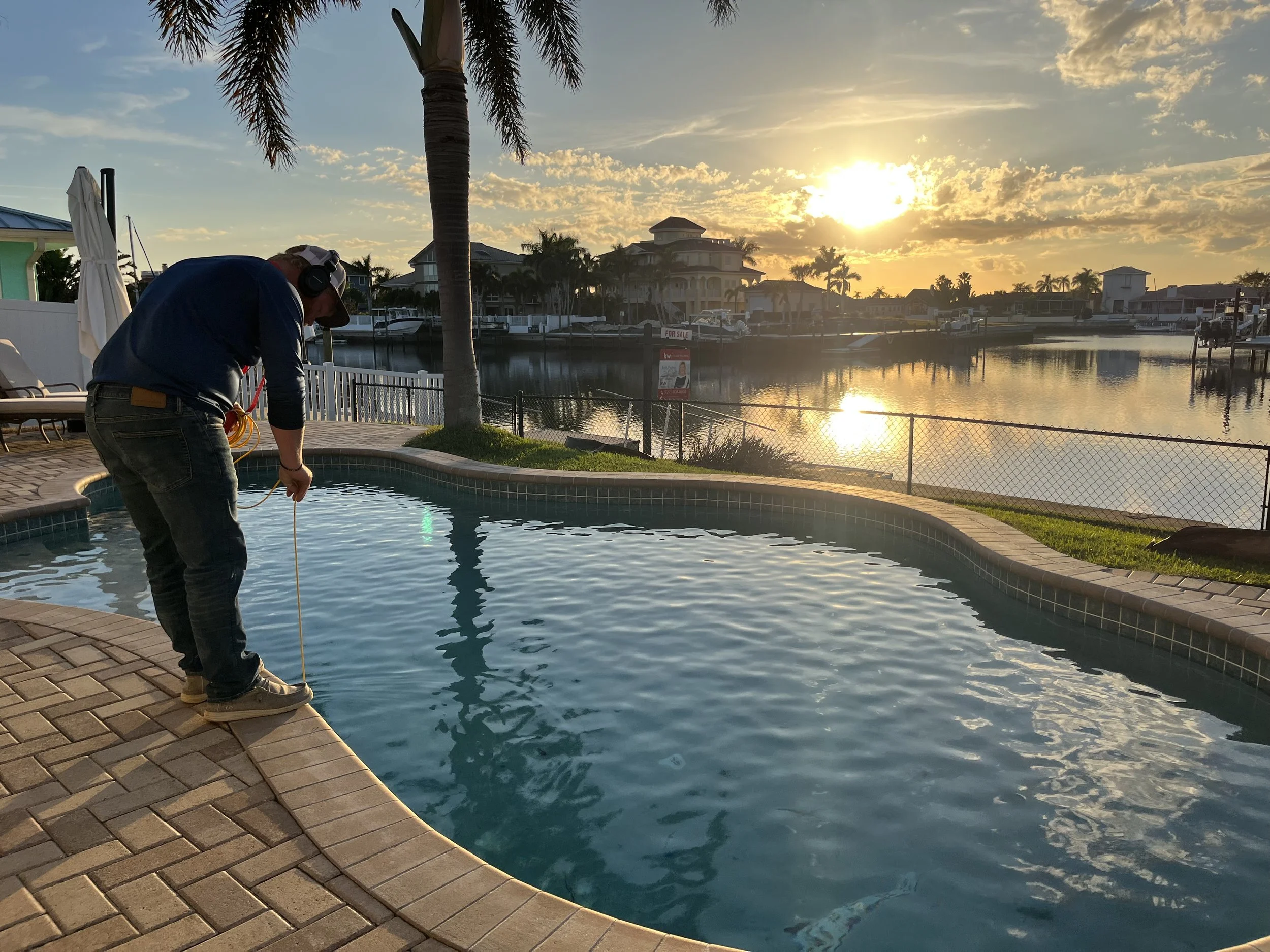 A man wearing a cap and headphones stands by the edge of a swimming pool, using a tool to check the water, with a scenic sunset over a body of water and houses in the background.