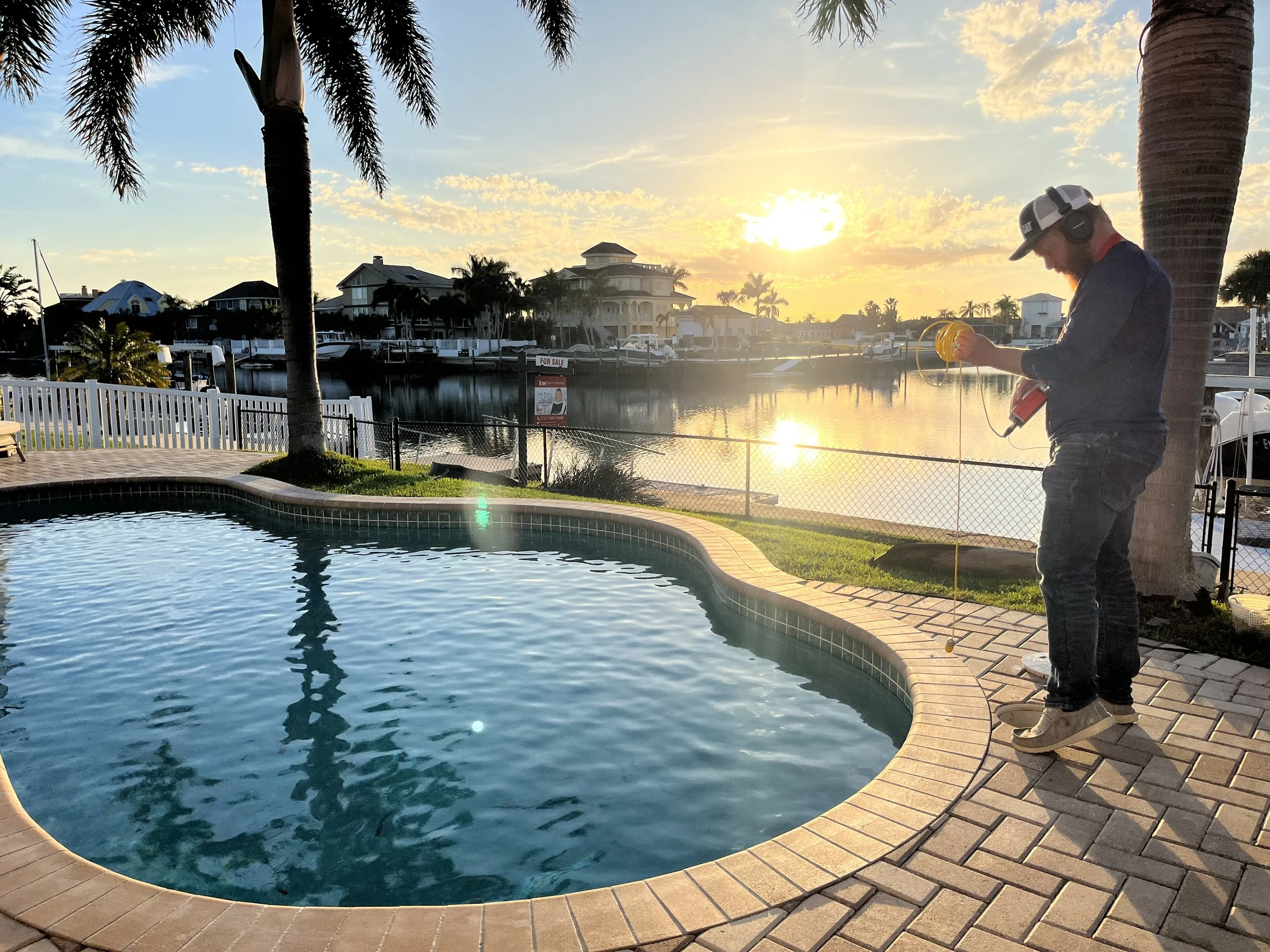 Man standing by a swimming pool holding a long tool with a reel, during sunset, with a canal, boats, and houses in the background.