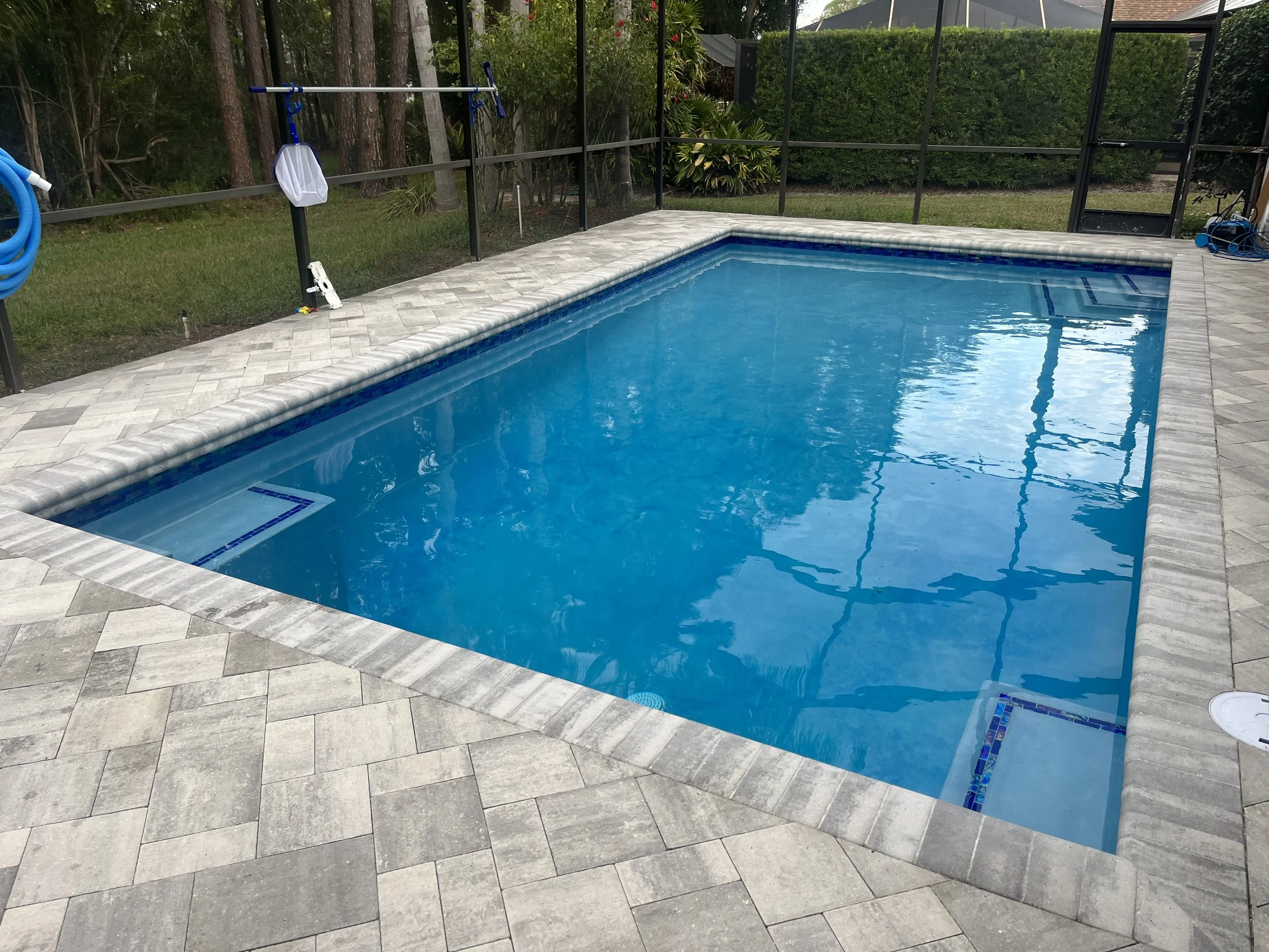 In-ground rectangular swimming pool with a paved stone deck, enclosed by a black metal fence, surrounded by trees and bushes, with ladders and pool cleaning equipment visible.
