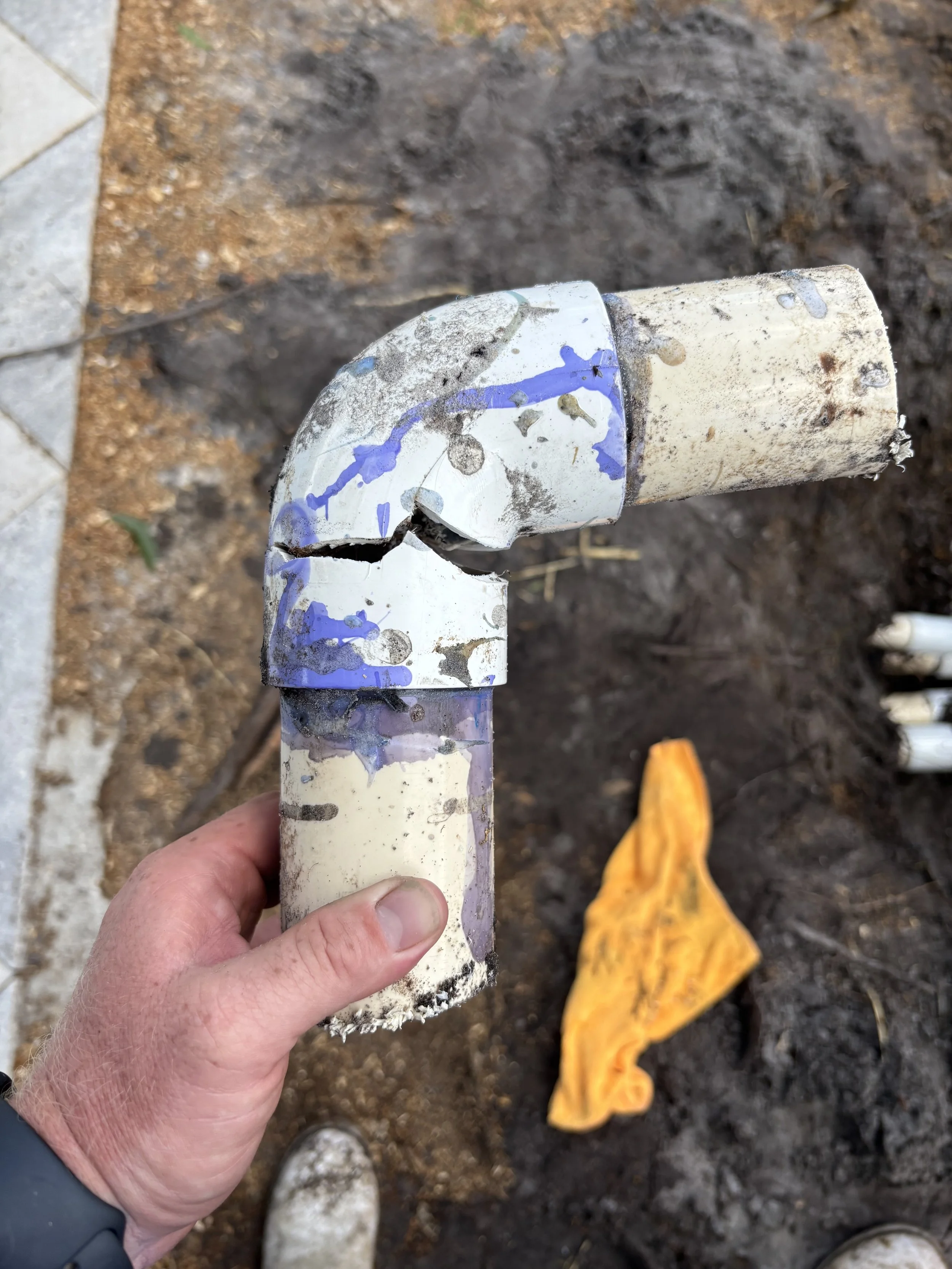 A person holding a damaged, cracked section of a white PVC pipe with blue paint splatters at a construction or repair site. The background shows dirt, a piece of orange cloth, and a concrete edge.