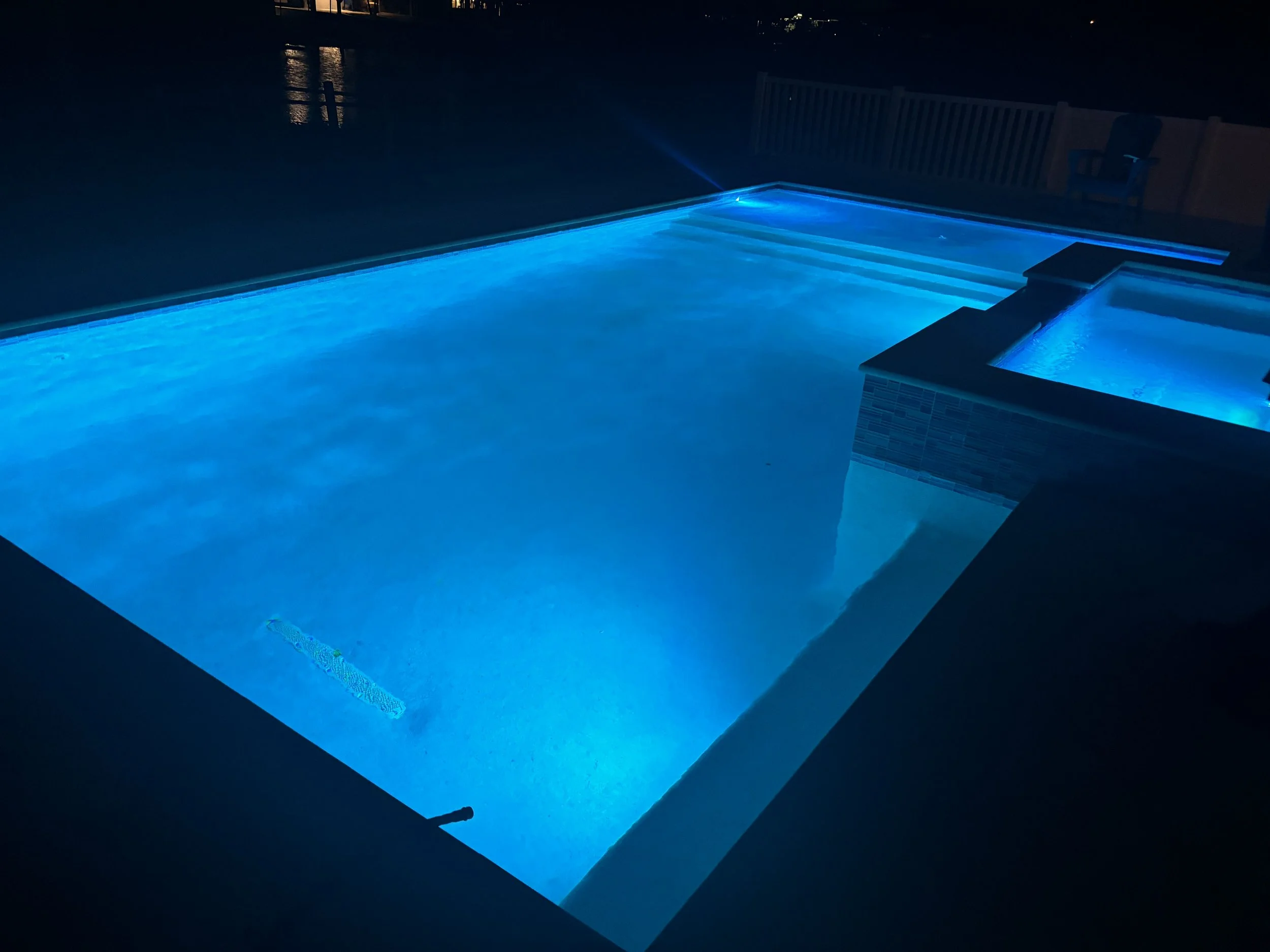 A lit swimming pool at night with blue lighting, a small hot tub attached, surrounded by a white fence, with reflections on the water surface.