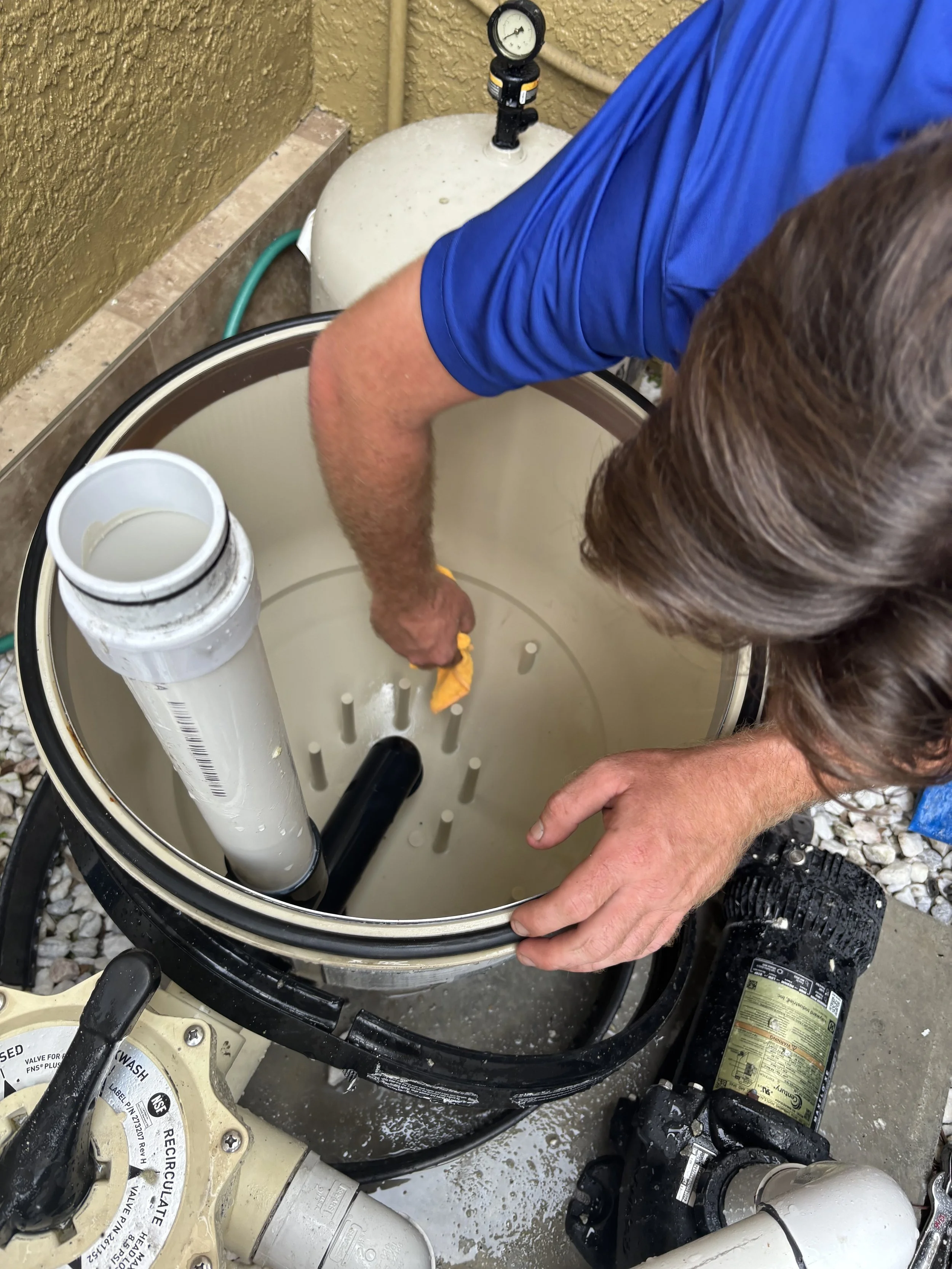 A person working inside a white cylindrical water filter housing, using a yellow cloth, with a pressure gauge on a white tank in the background and plumbing connections around.