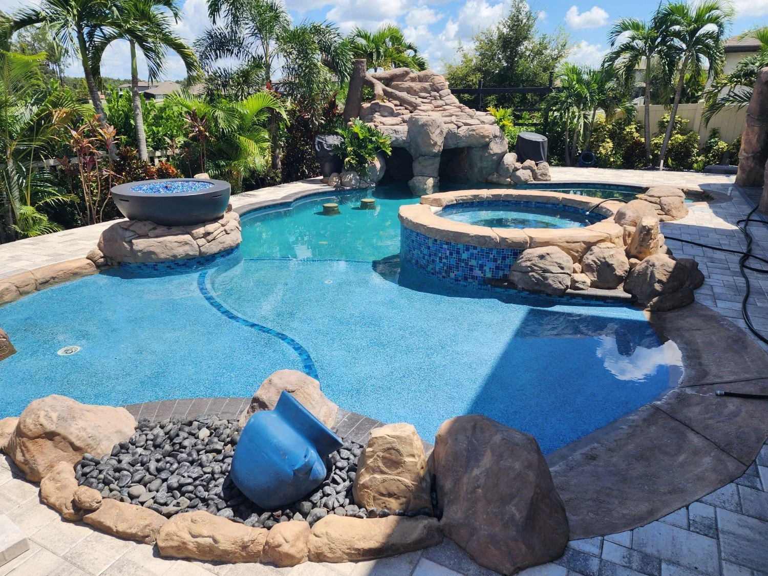 A backyard swimming pool and hot tub surrounded by rocks, palm trees, and lush greenery, with a stone waterfall feature in the background.
