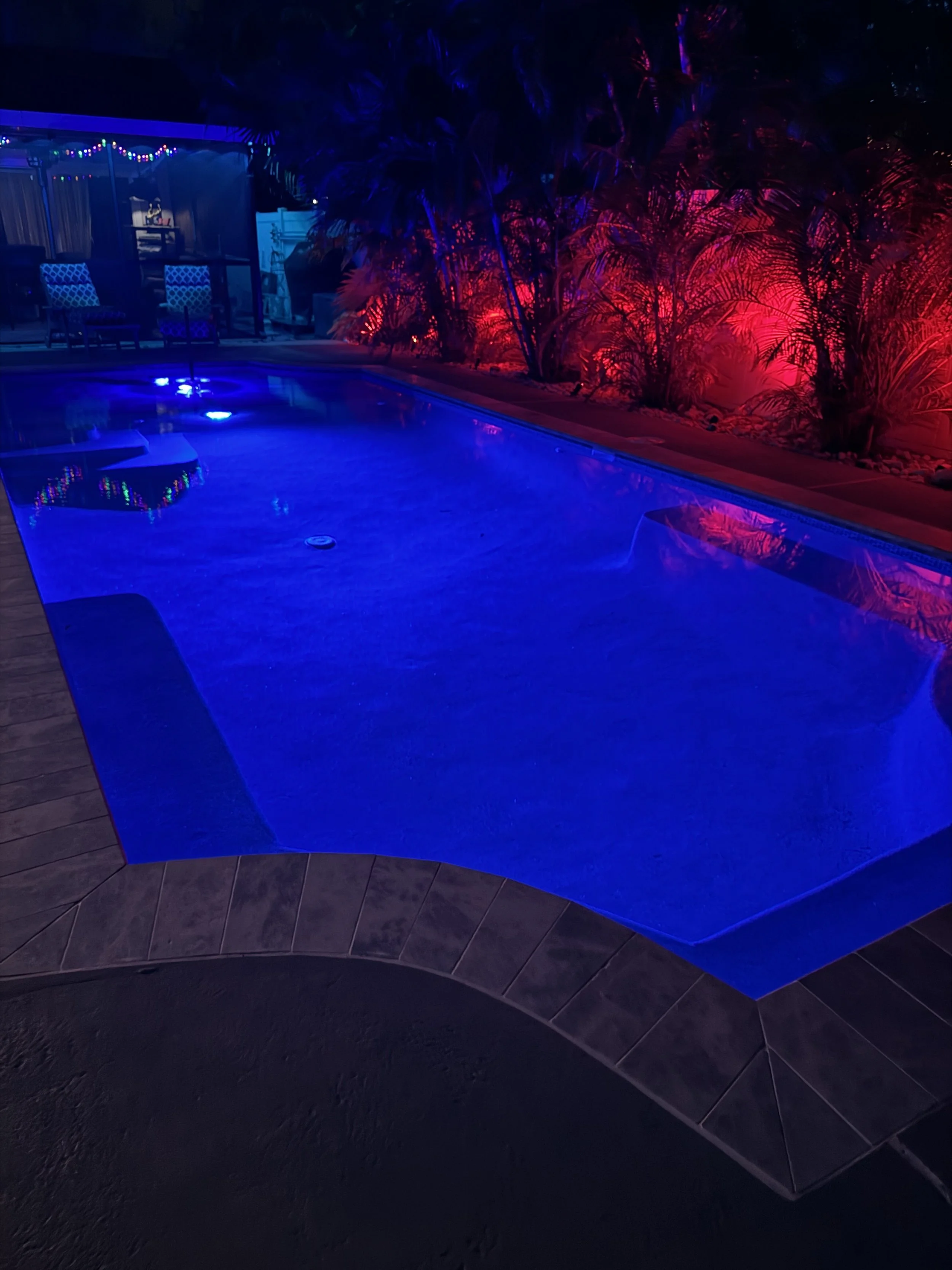 A swimming pool illuminated with vibrant blue and red lighting at night, with surrounding lush tropical plants and a seating area with chairs and string lights in the background.
