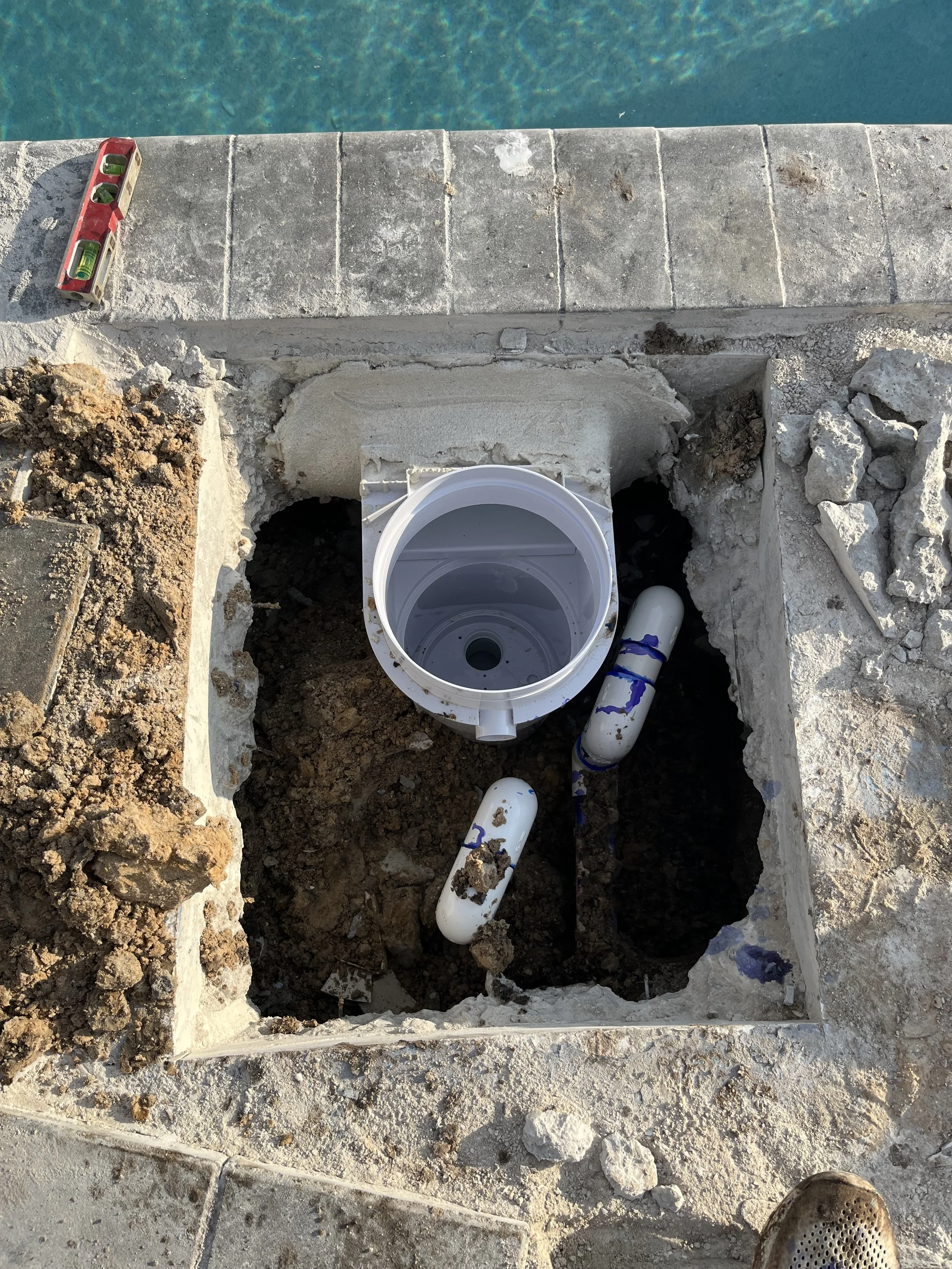 Underground plumbing installation in progress with open trench, PVC pipes, and a white cylindrical fittings, near a stone patio overlooking a pool.