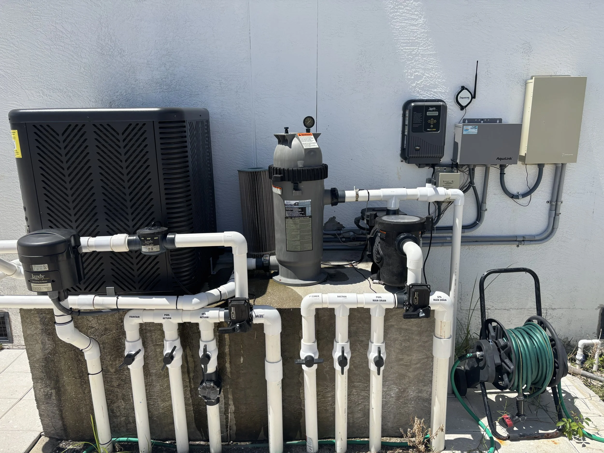A pool filtration and pump system installed outside on a concrete surface against a white stucco wall, with various pipes, valves, and electrical equipment, and a green garden hose on a reel.